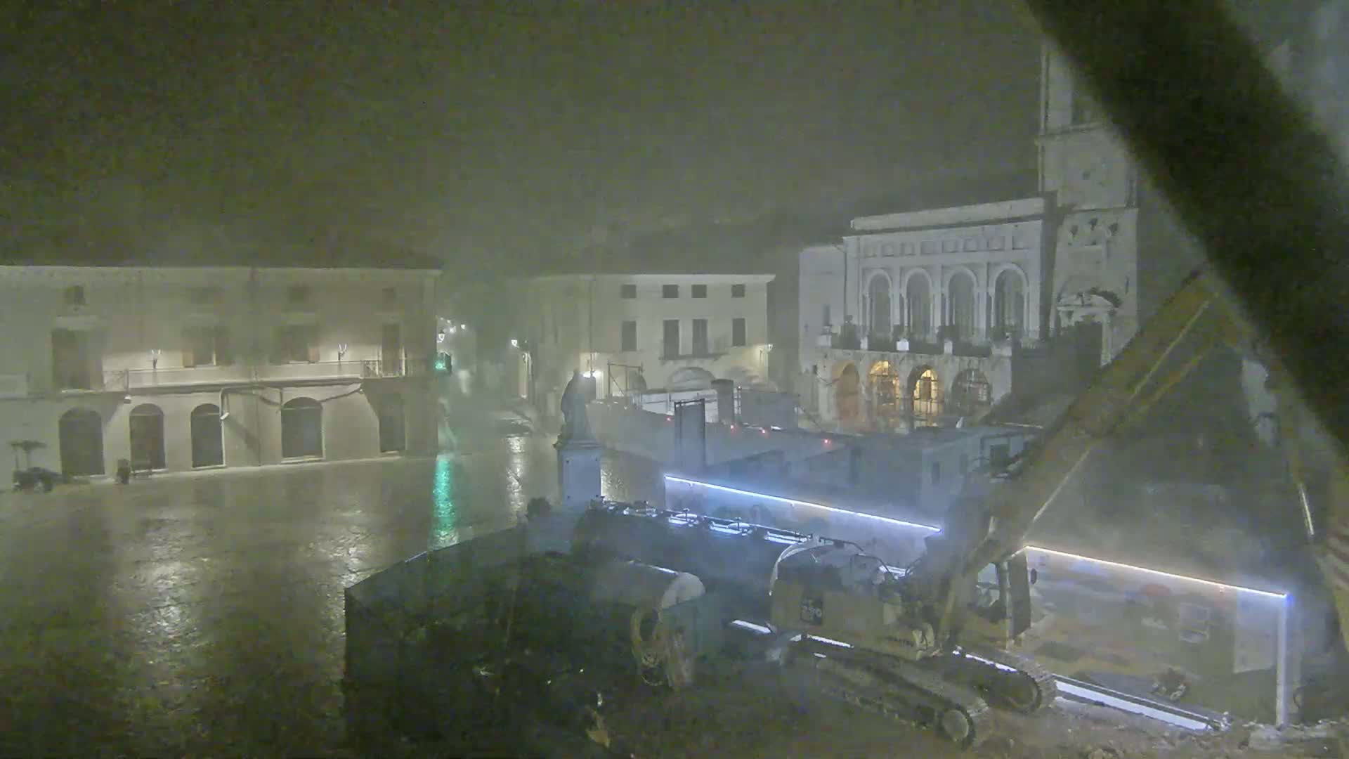 A nighttime view of a rain-soaked town square shows construction equipment near several buildings, one of which appears to be a church.