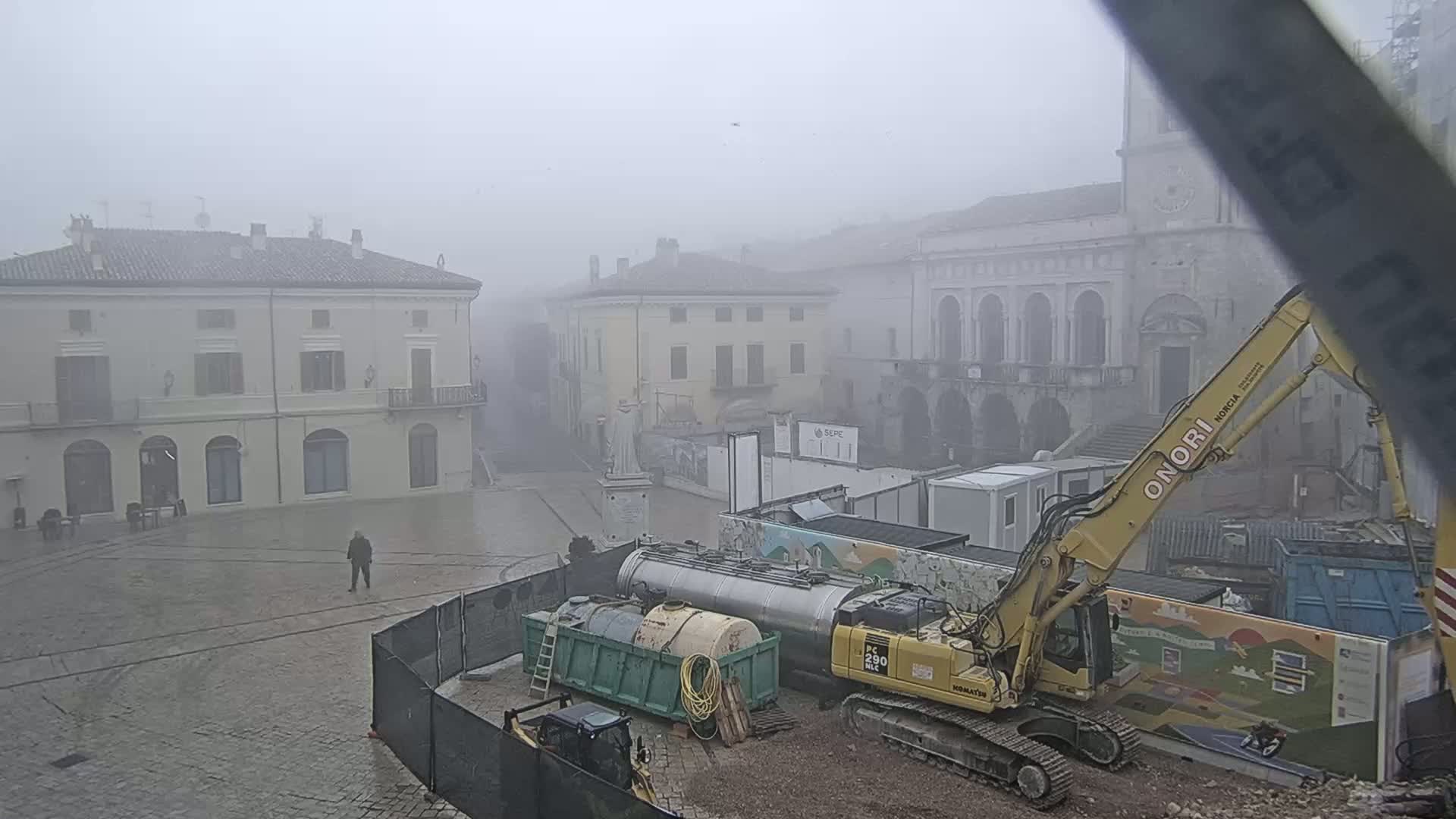 A foggy town square with several buildings, construction equipment including a large excavator, and a single person walking.