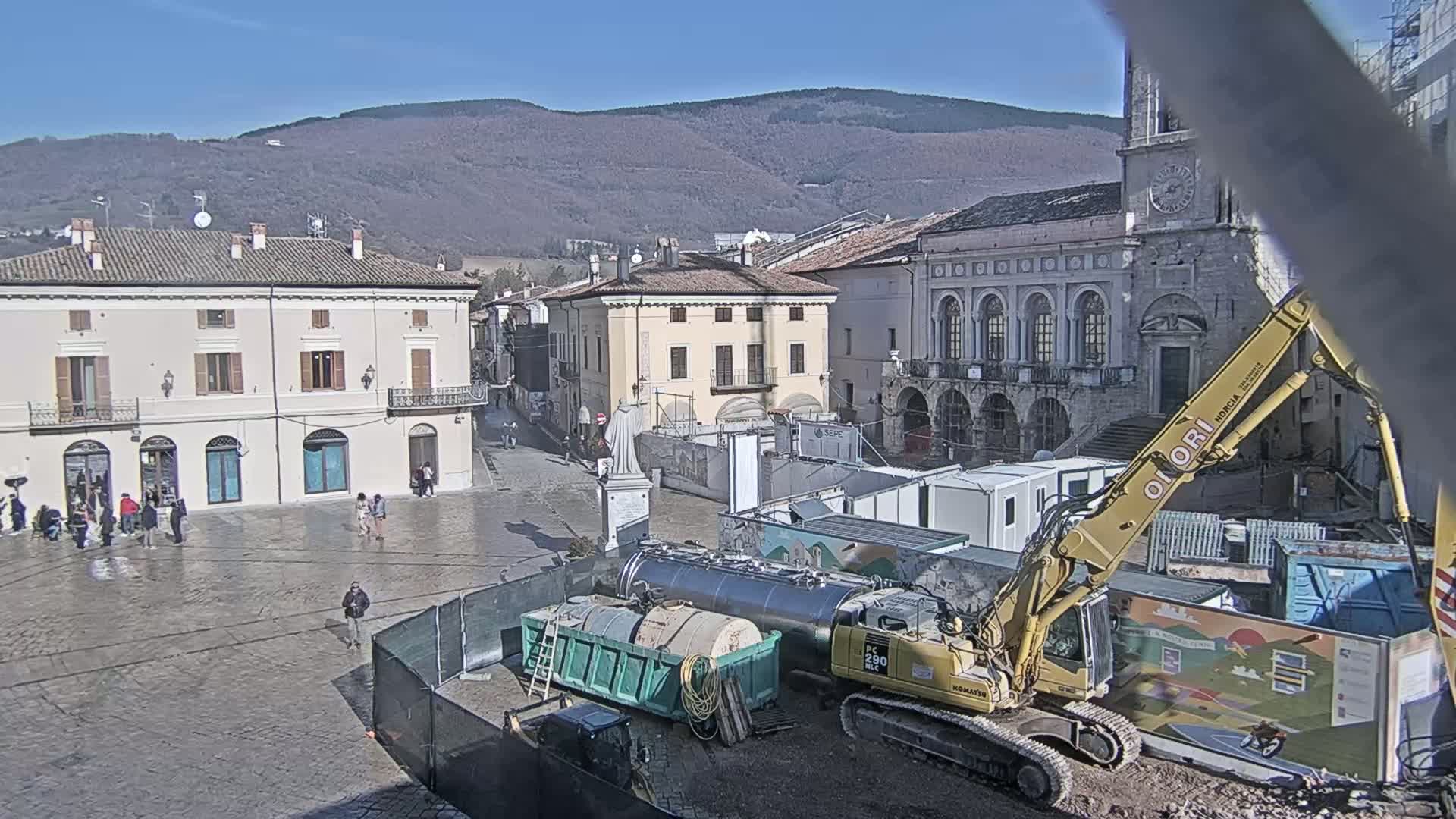 A town square under construction on a sunny day, with mountains in the background, features several buildings, construction equipment including an excavator and dumpsters, and a few people.