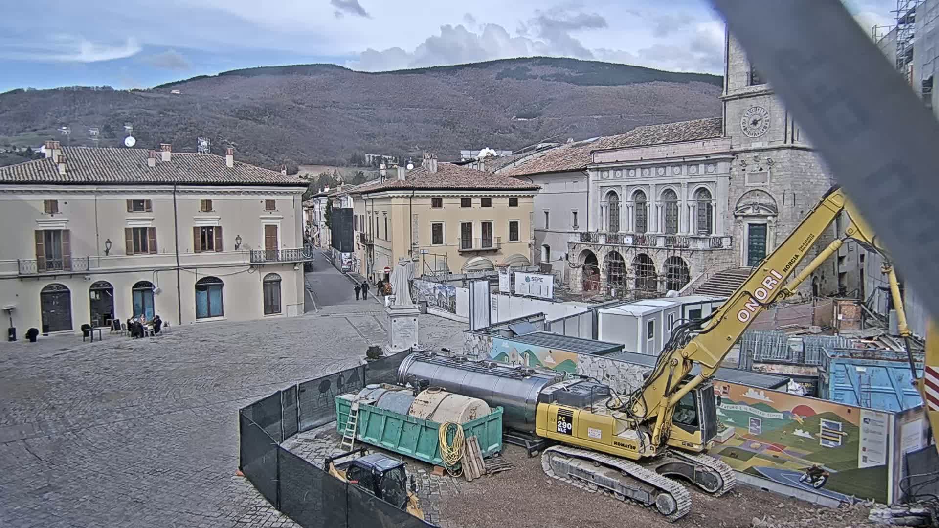 A town square under cloudy skies is undergoing construction, with an excavator and other equipment present near several buildings, one of which is a large, ornate stone structure.