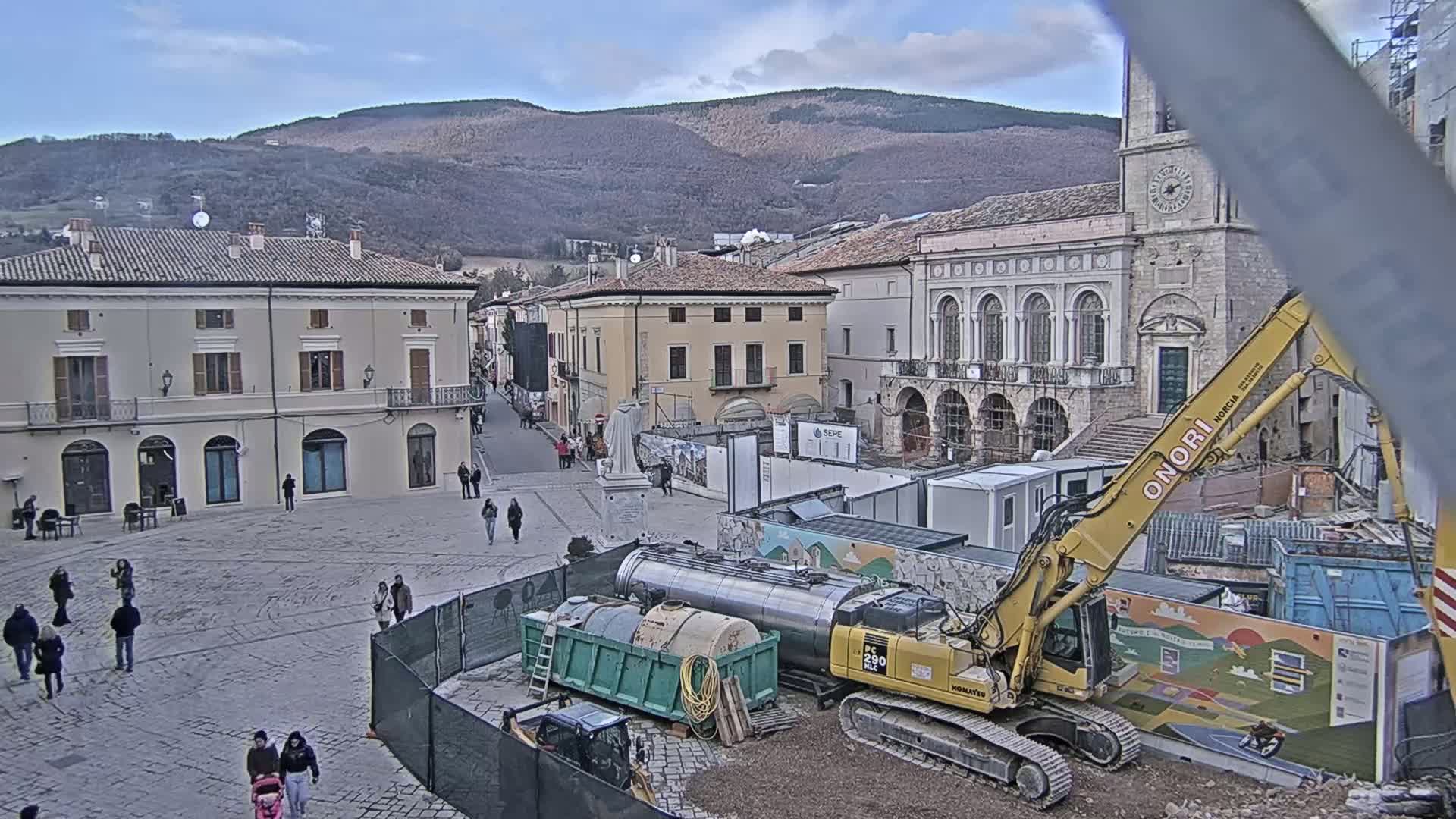 A town square under construction on a partly cloudy day, with mountains in the background, shows construction equipment and workers amidst several buildings.