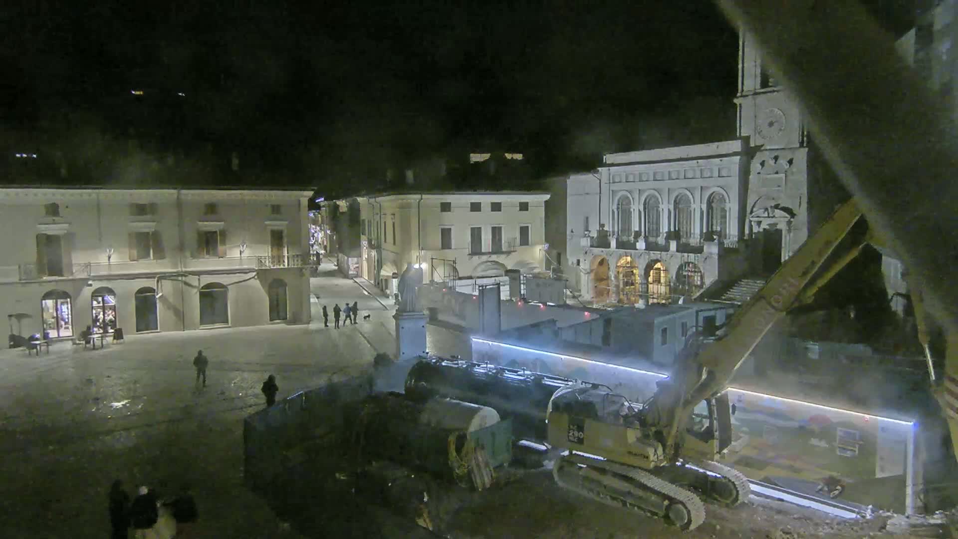 A nighttime view of a town square under construction, showing several buildings, construction equipment, and a few people.