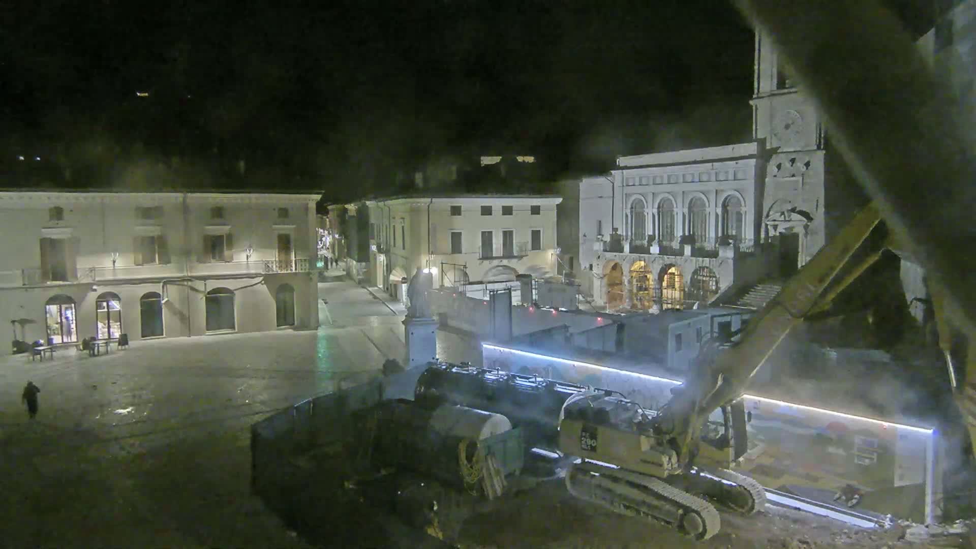 At night, construction equipment is working in a town square near several buildings.