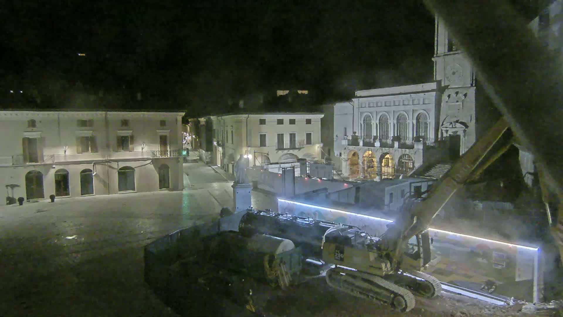 At night, construction equipment operates in a town square near classical-style buildings.