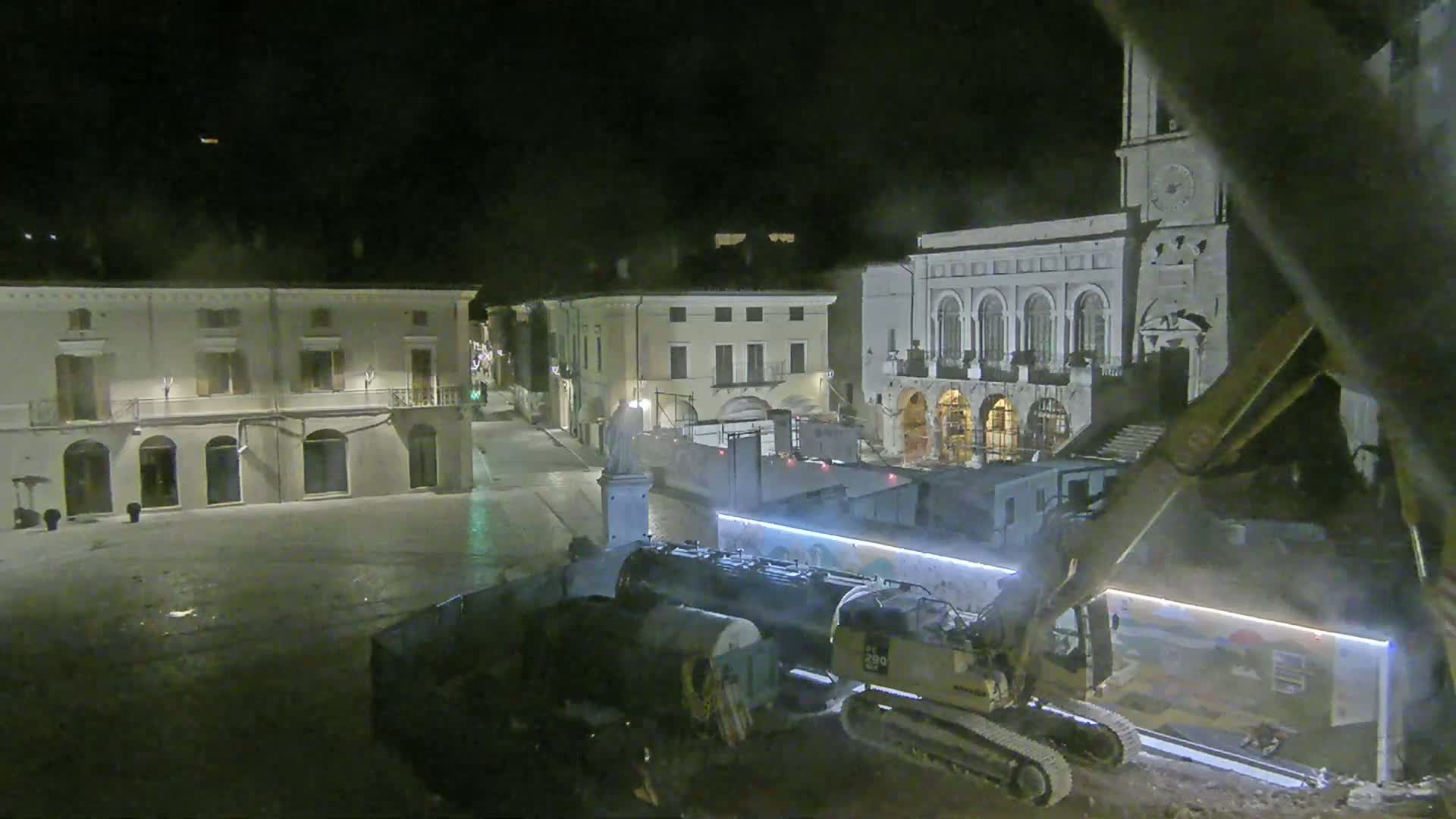 At night, heavy machinery works on a town square next to several buildings, one of which has a clock tower.