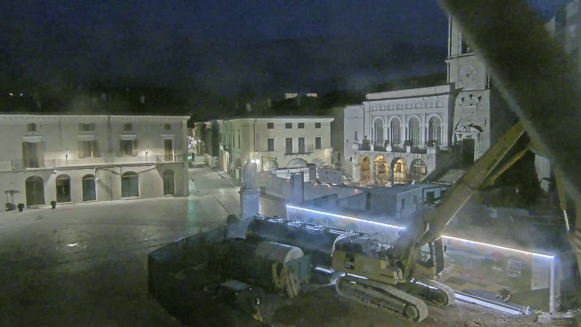 At night, construction equipment sits in a town square in front of several buildings, one of which has a clock tower.