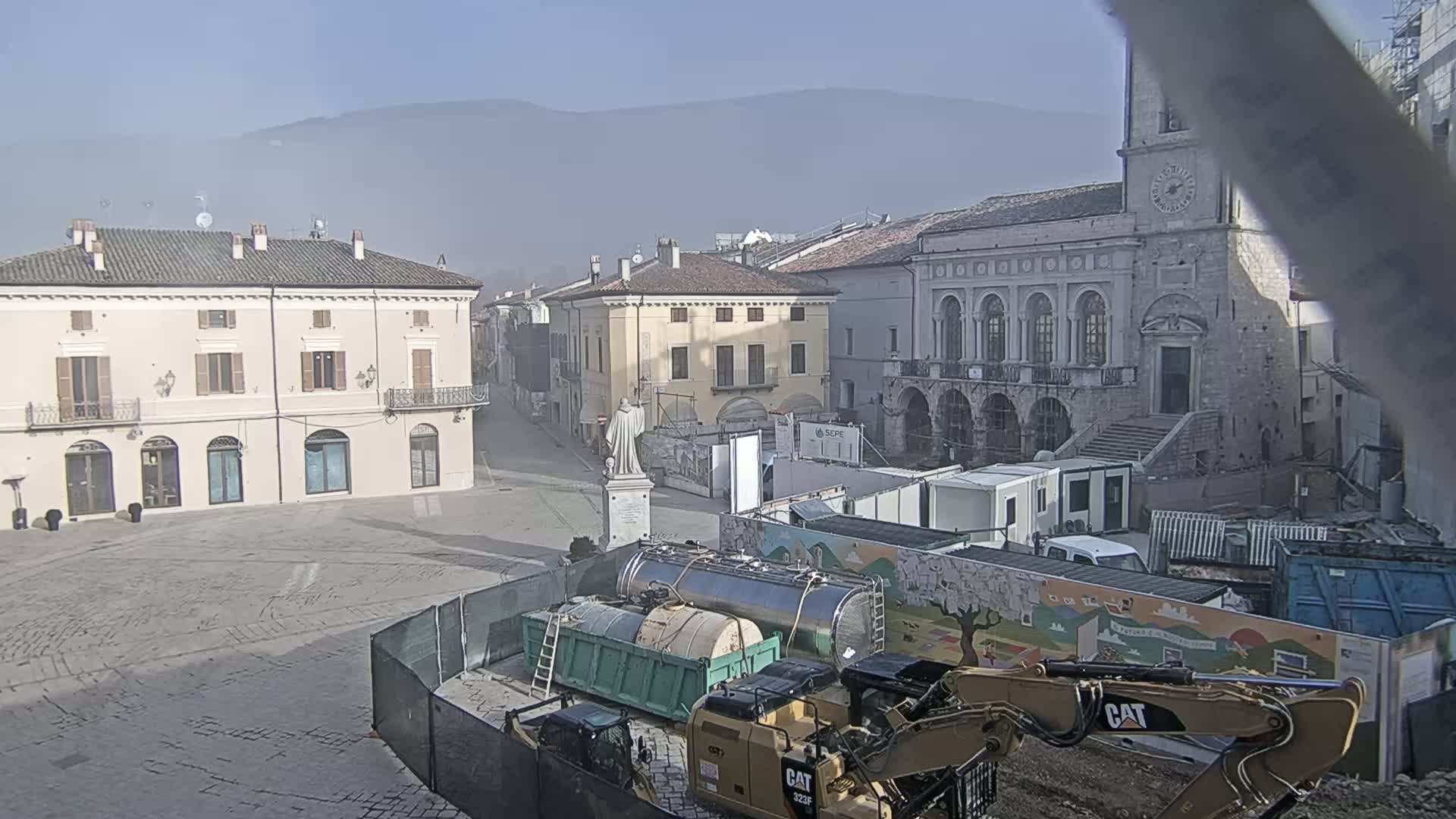 A mostly empty town square is under construction, with several pieces of heavy machinery and construction containers present, on a hazy, sunny day with mountains in the background.