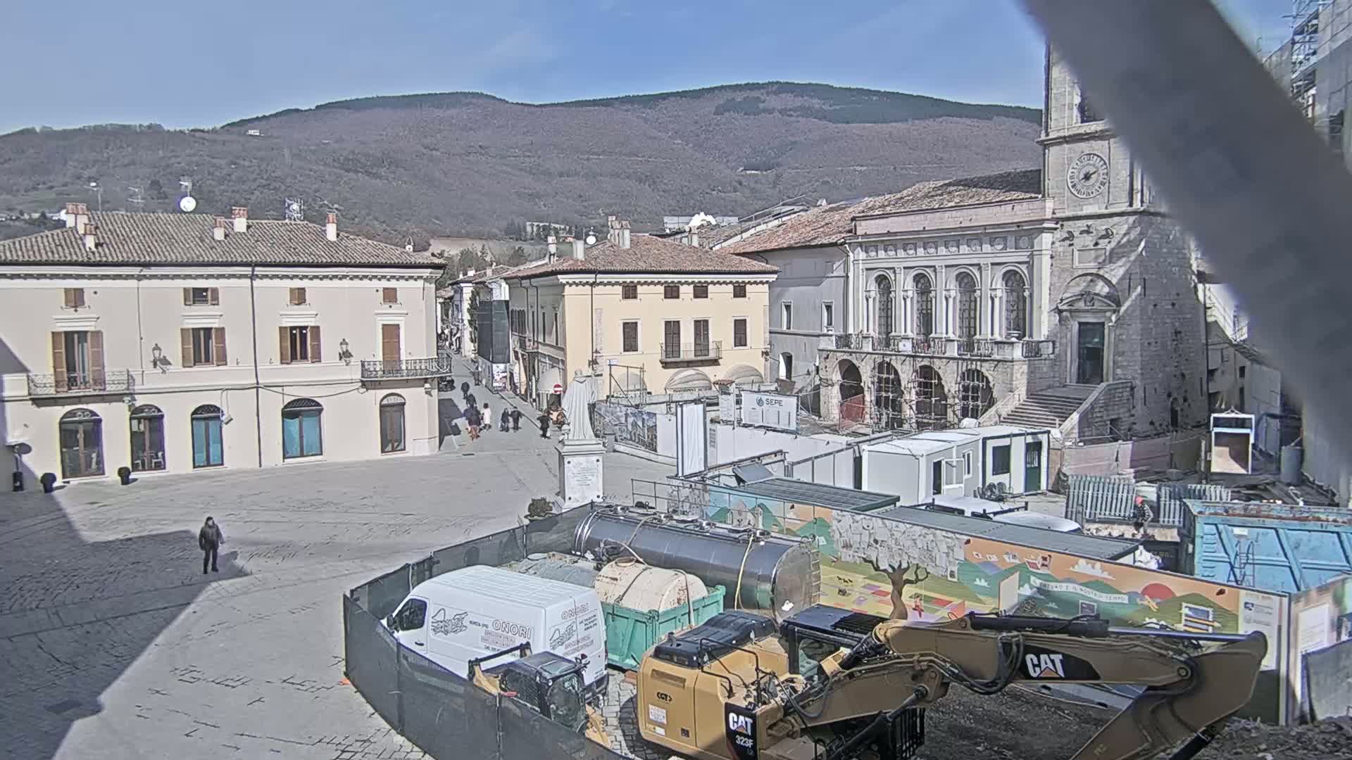 A town square under construction on a sunny day, with several buildings surrounding it, construction equipment, and a few people walking around.