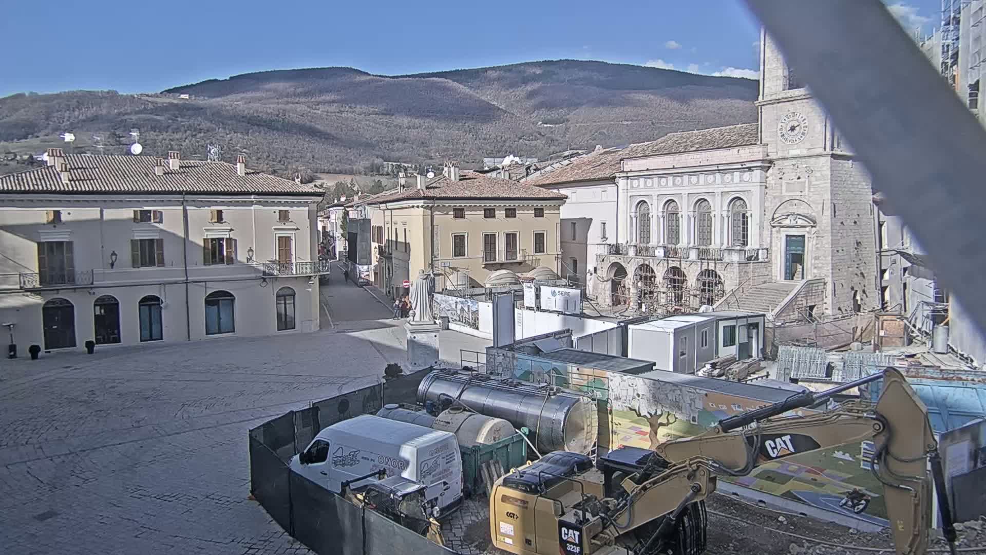 A sunny day reveals a town square under construction, with several buildings visible, including a large stone building with arched windows, and construction equipment, including a backhoe and tankers, in the foreground.