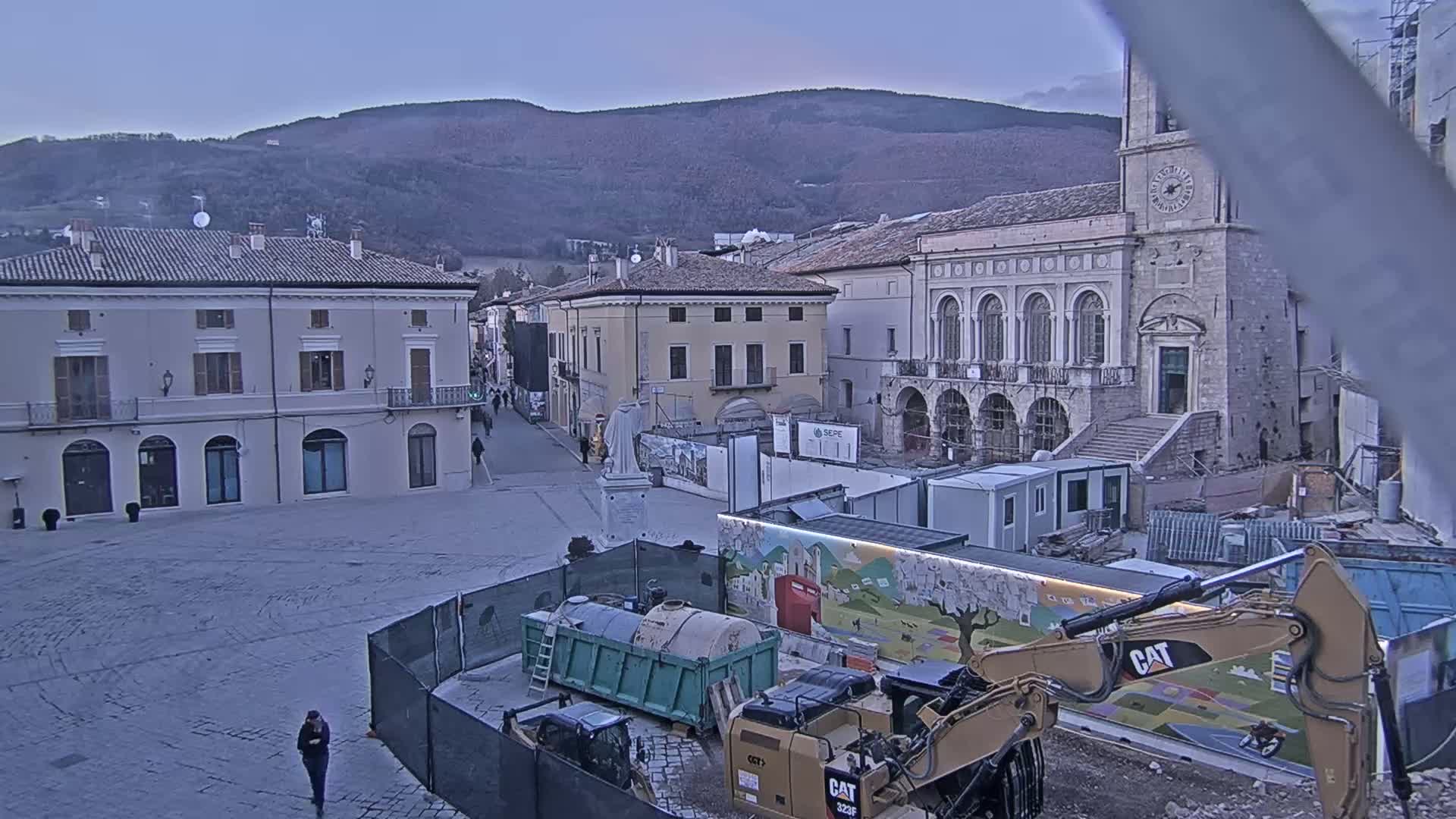 A town square under construction, featuring heavy machinery, temporary structures, and older buildings against a backdrop of mountains under a dusky sky.