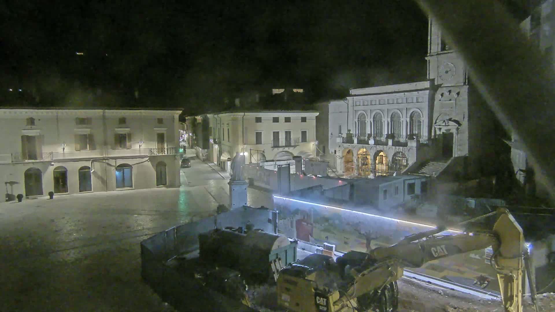 At night, construction equipment operates in a town square near classical-style buildings.