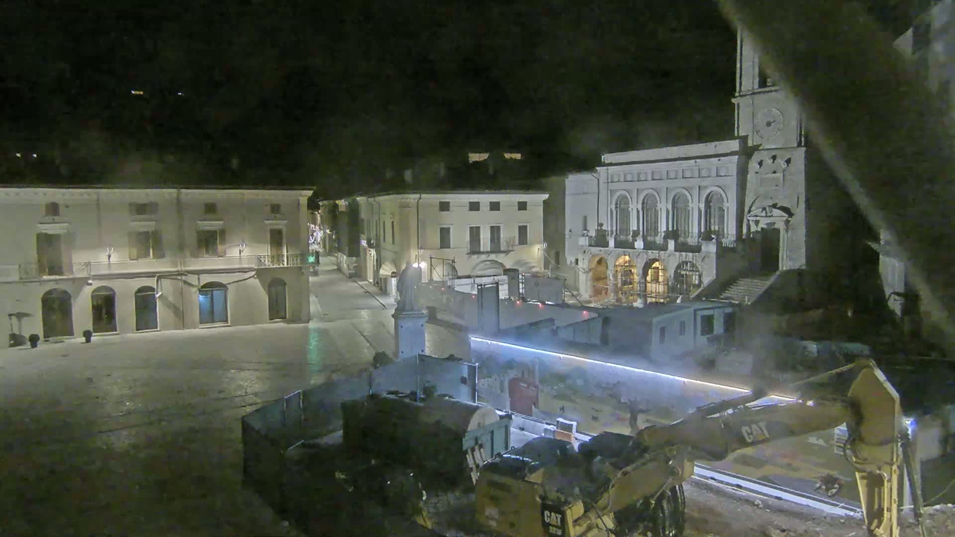 At night, construction equipment sits in a town square near several buildings, one of which is a large structure with arched windows and a clock tower.