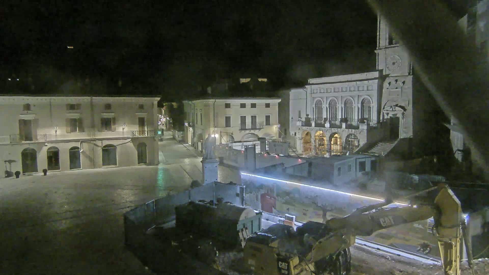 At night, construction equipment is working in a town square next to several buildings, one of which is a large building with a clock tower.