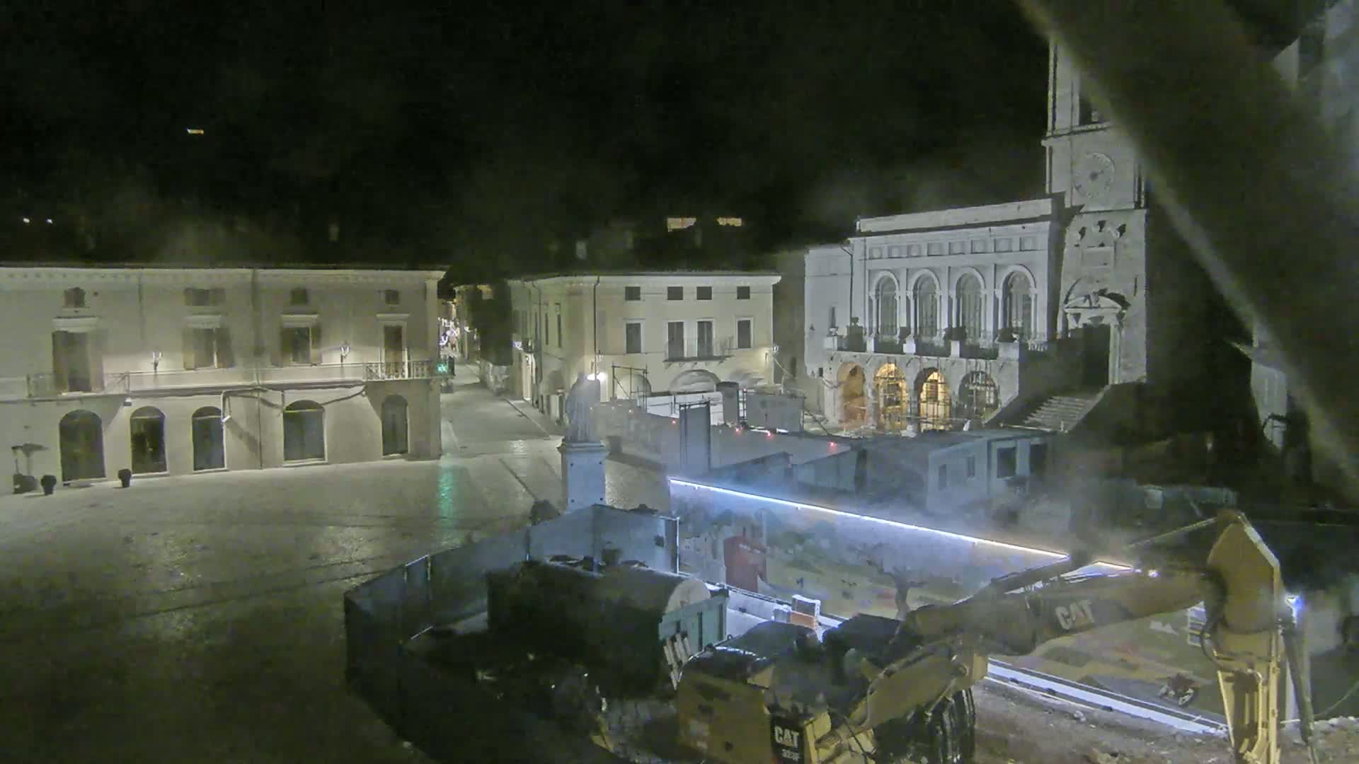 At night, construction equipment sits in a town square surrounded by old buildings under a dark sky.