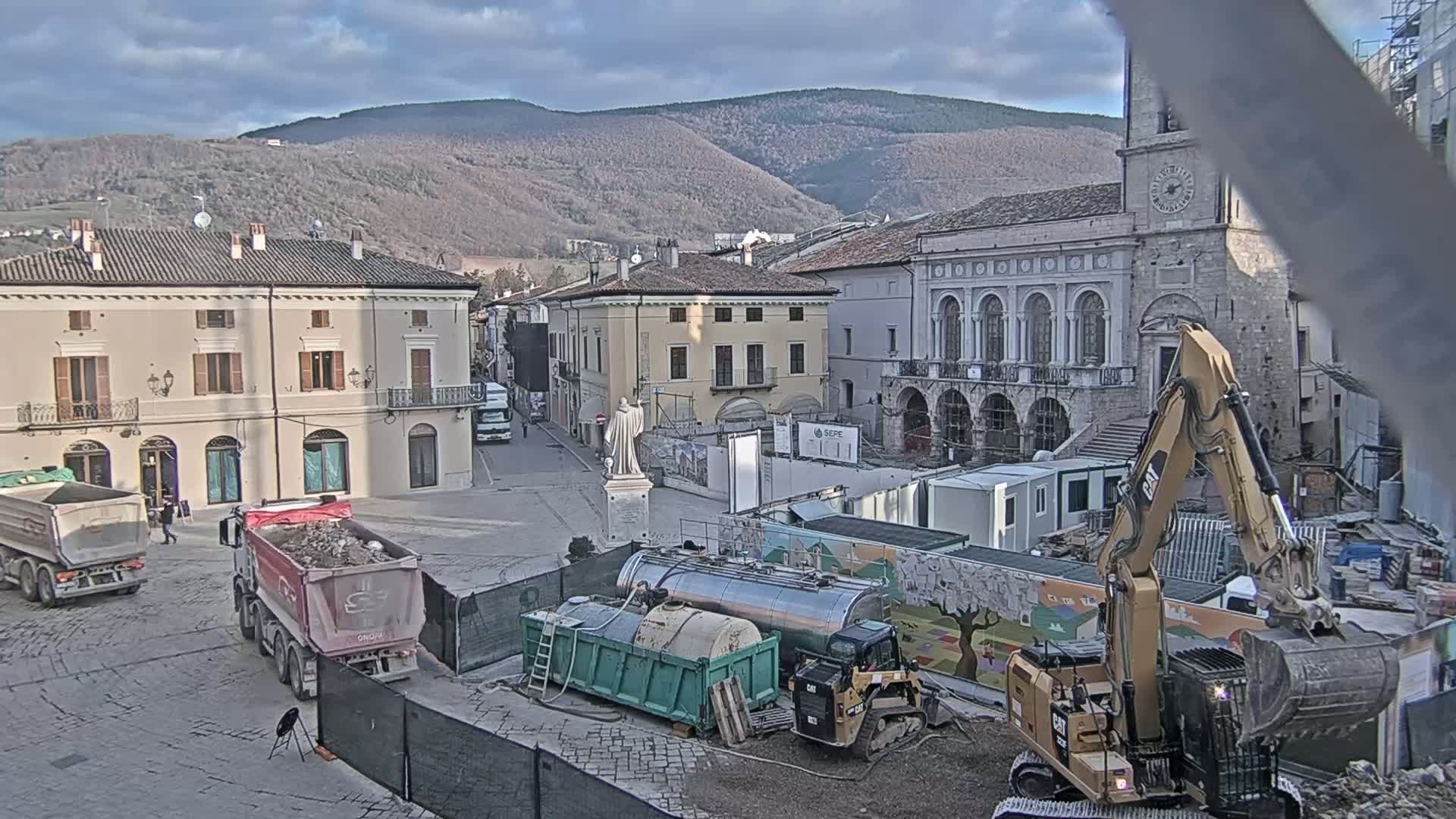 A town square under construction, with dump trucks, heavy machinery, and buildings under renovation, is situated against a backdrop of mountains under a partly cloudy sky.