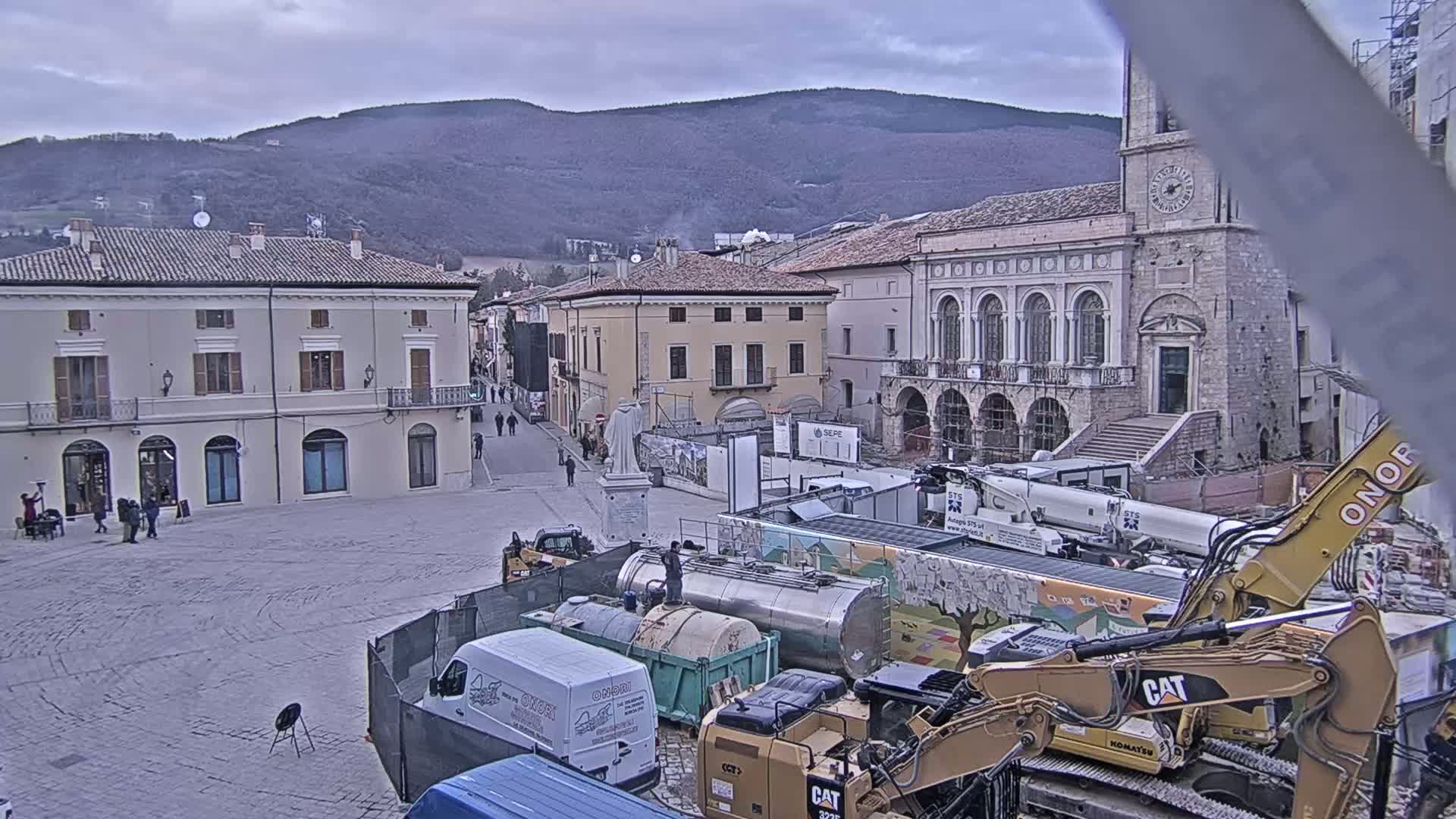 A town square under an overcast sky is undergoing construction, with several pieces of heavy machinery and construction vehicles present among older buildings.