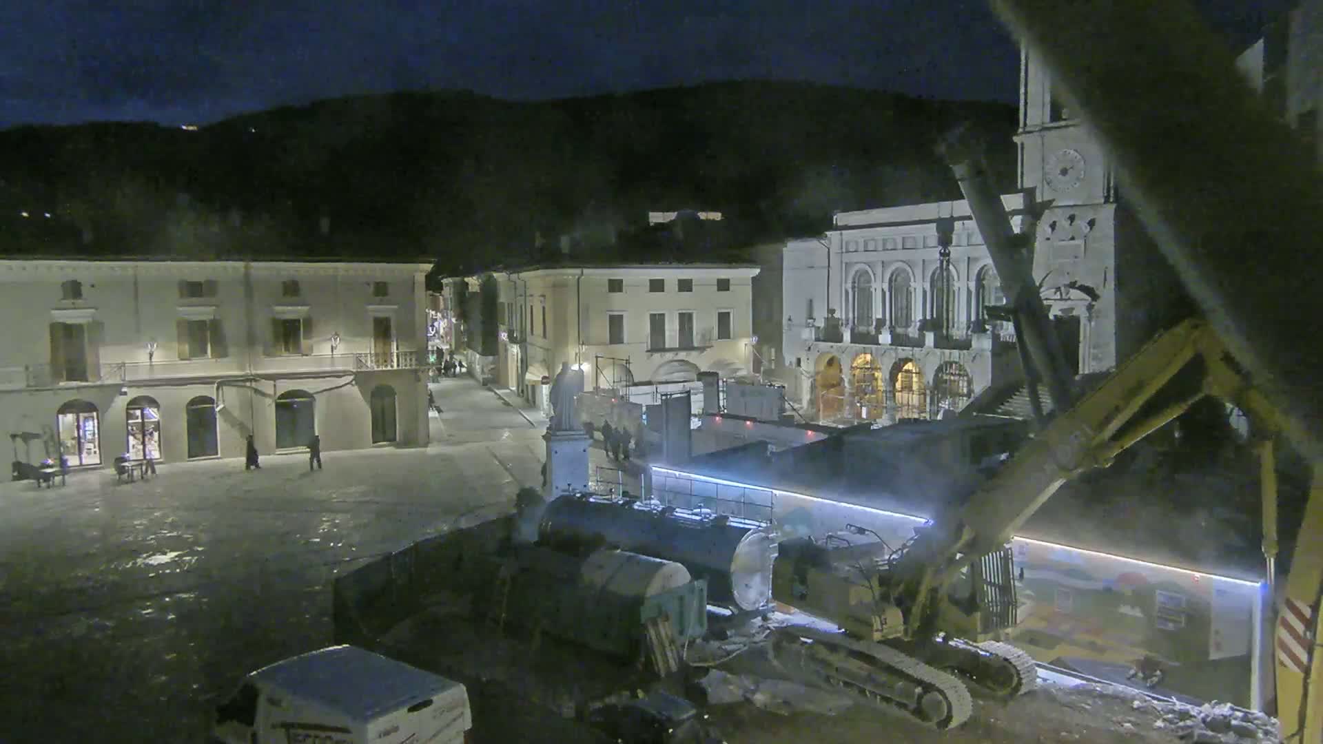 At night, construction equipment operates in a town square near buildings, under a dark sky.