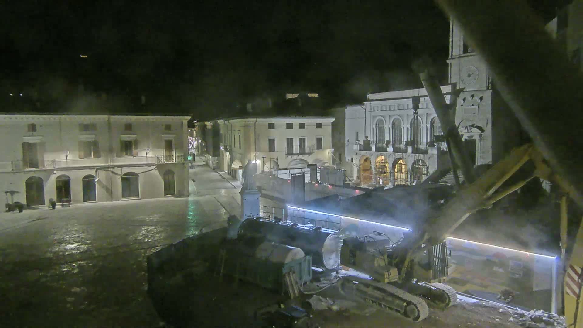 At night, construction equipment works in a town square surrounded by buildings under a hazy, possibly snowy or misty sky.
