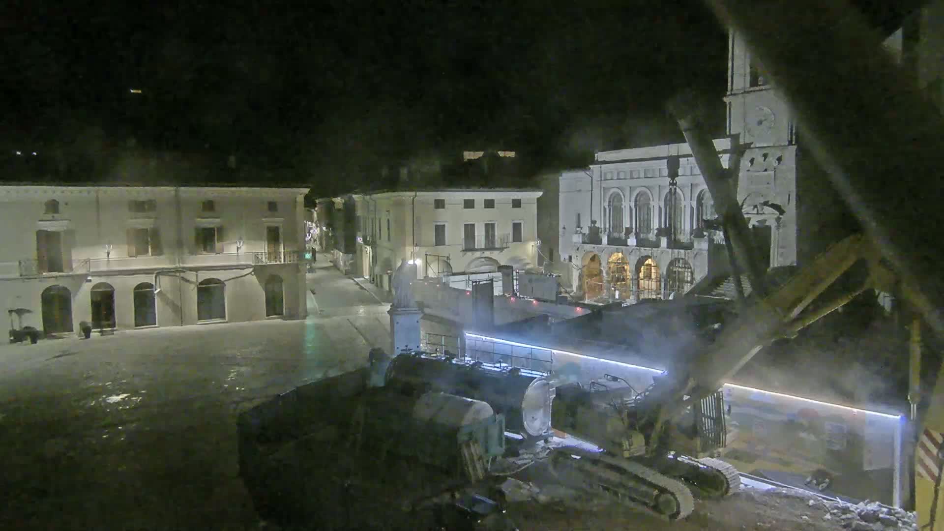 At night, construction equipment is working in a town square surrounded by buildings, under hazy conditions.
