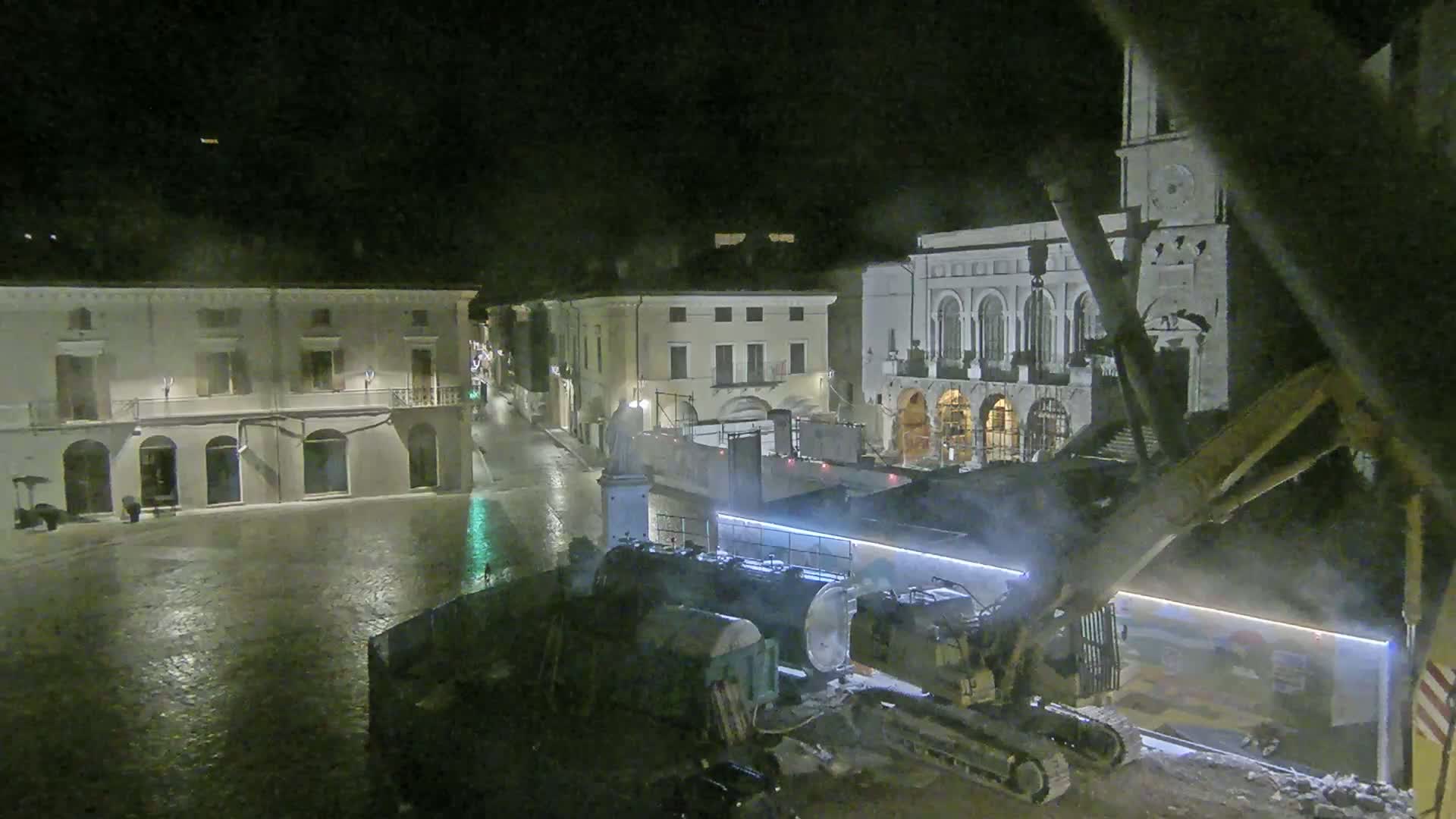 At night, under rainy conditions, heavy machinery is working in a town square next to historic buildings.