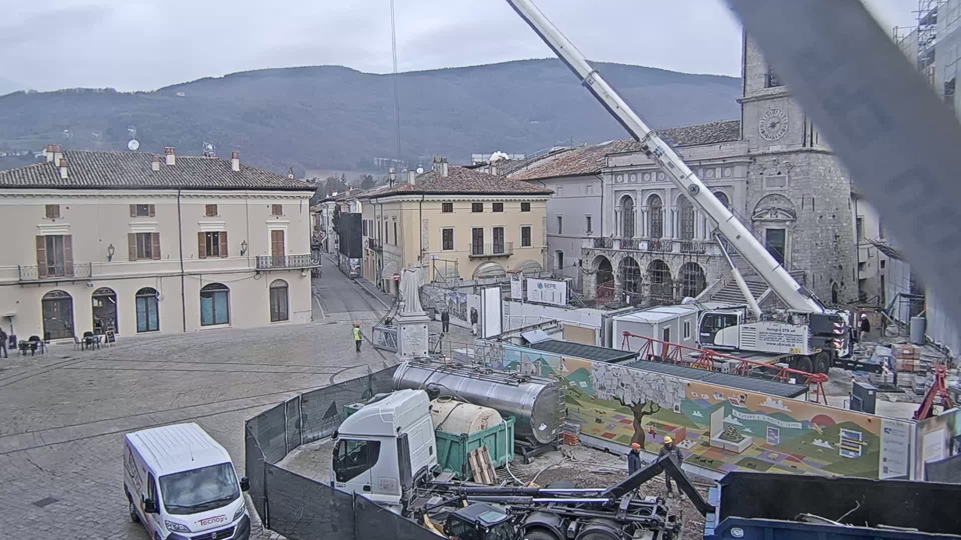 A town square under a cloudy sky is undergoing construction, with a large crane, trucks, and workers present.