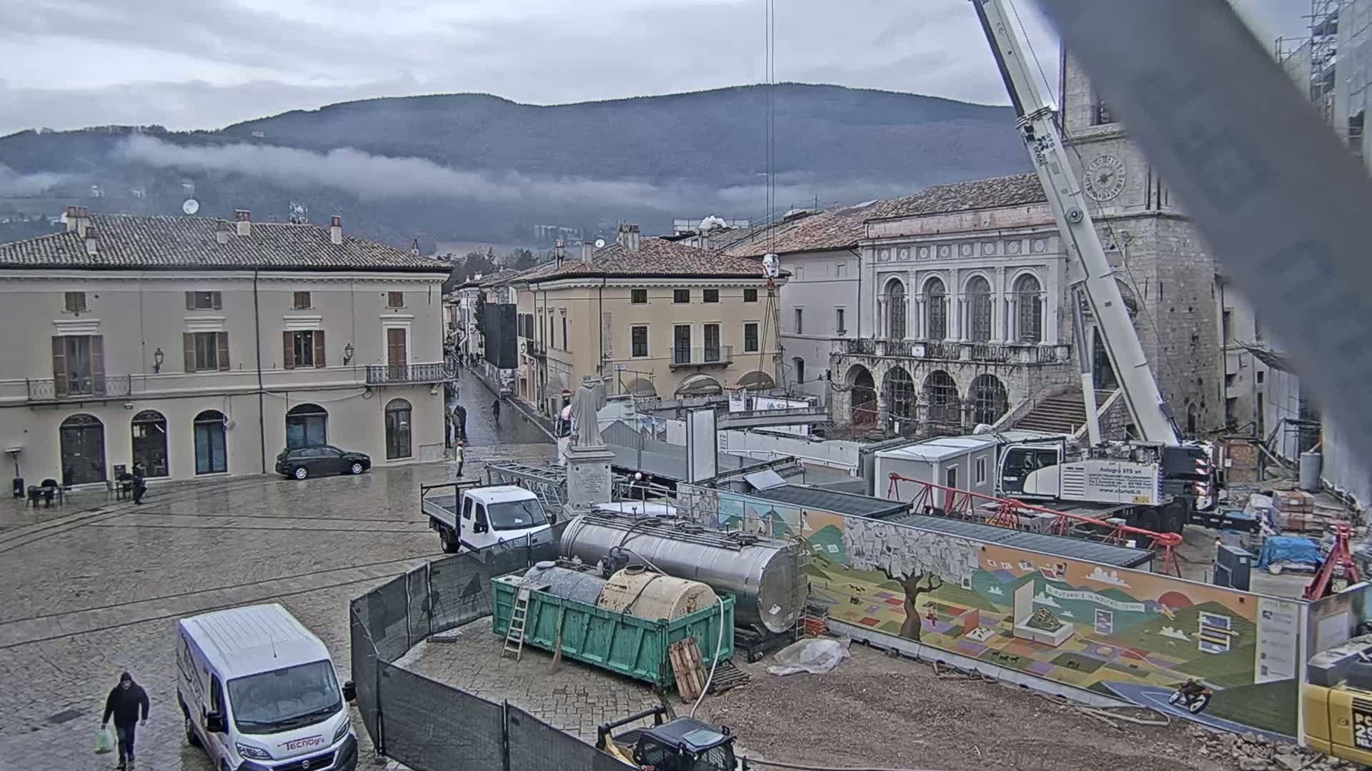 A town square under a cloudy sky is undergoing construction, with several trucks, a crane, and building materials present.