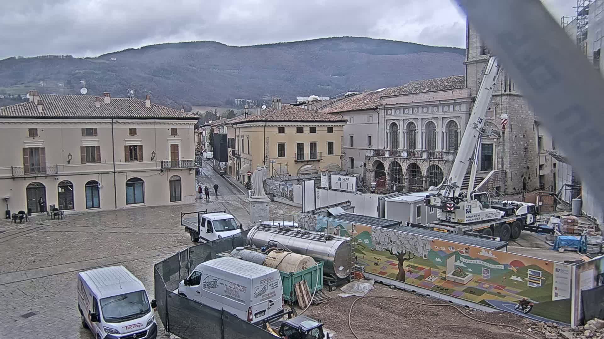 A town square under an overcast sky features construction equipment, vans, and older buildings, with mountains visible in the background.