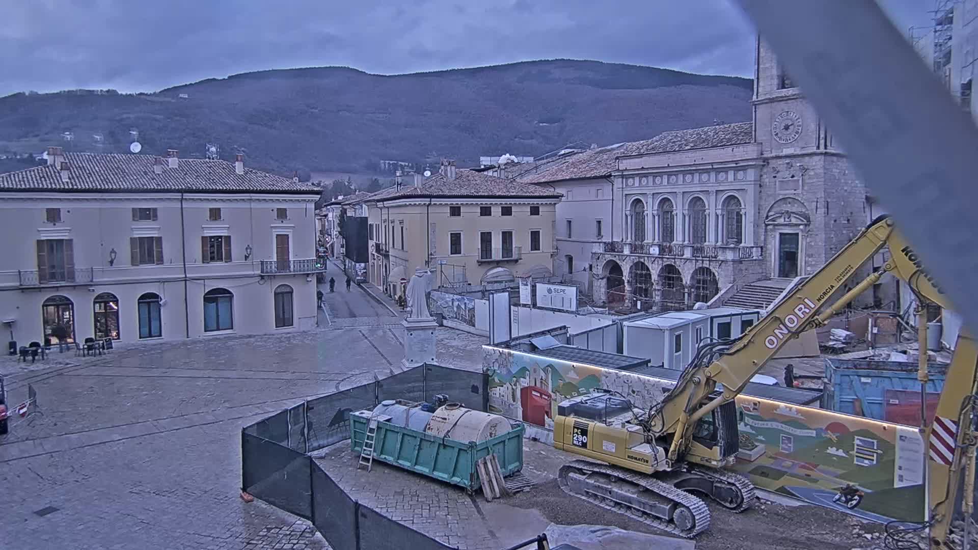 A town square under a cloudy sky is undergoing construction, with an excavator and other equipment present near classical-style buildings and mountains in the background.
