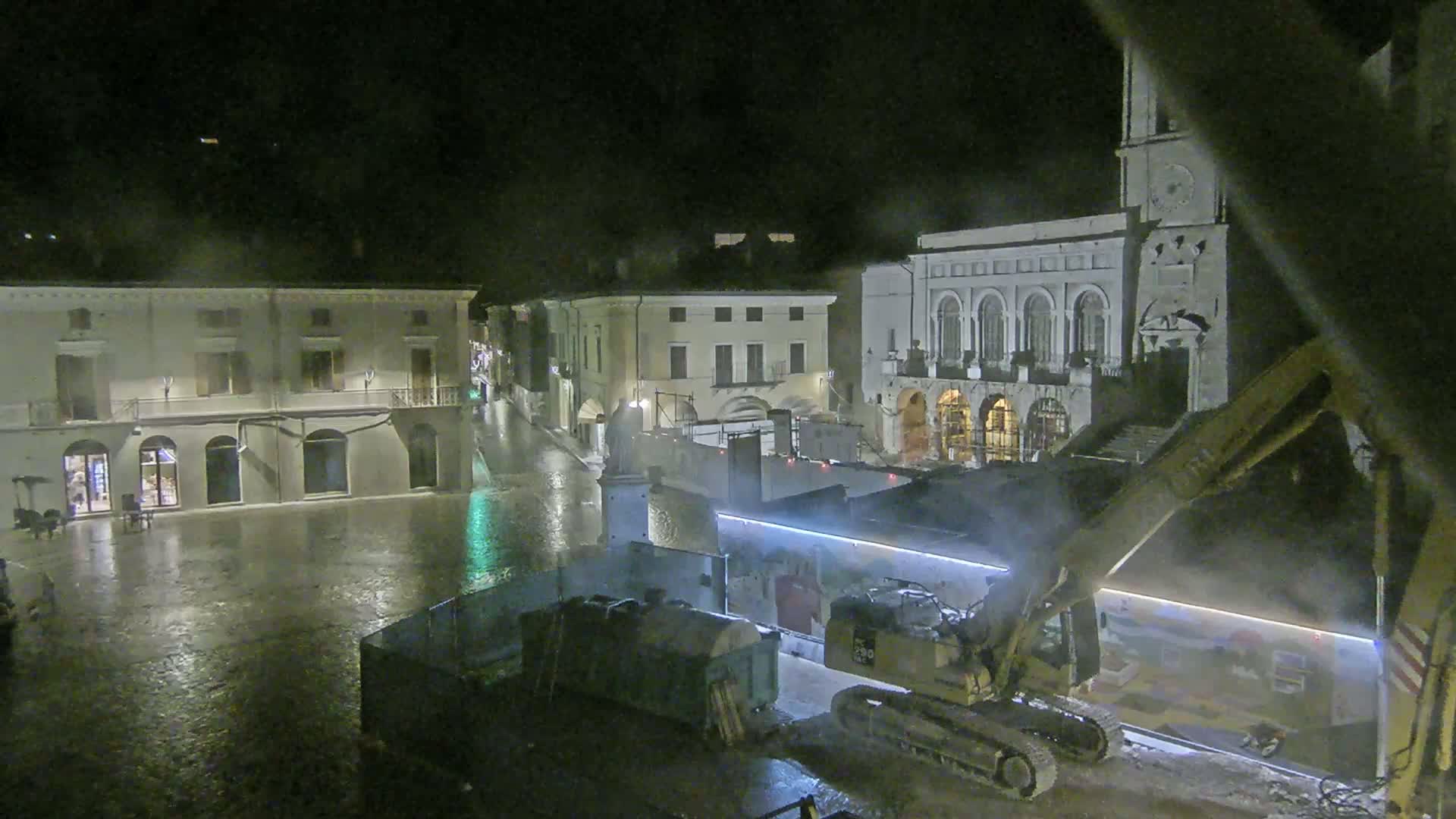 A nighttime view of a town square under rain, showing construction equipment near classical-style buildings.