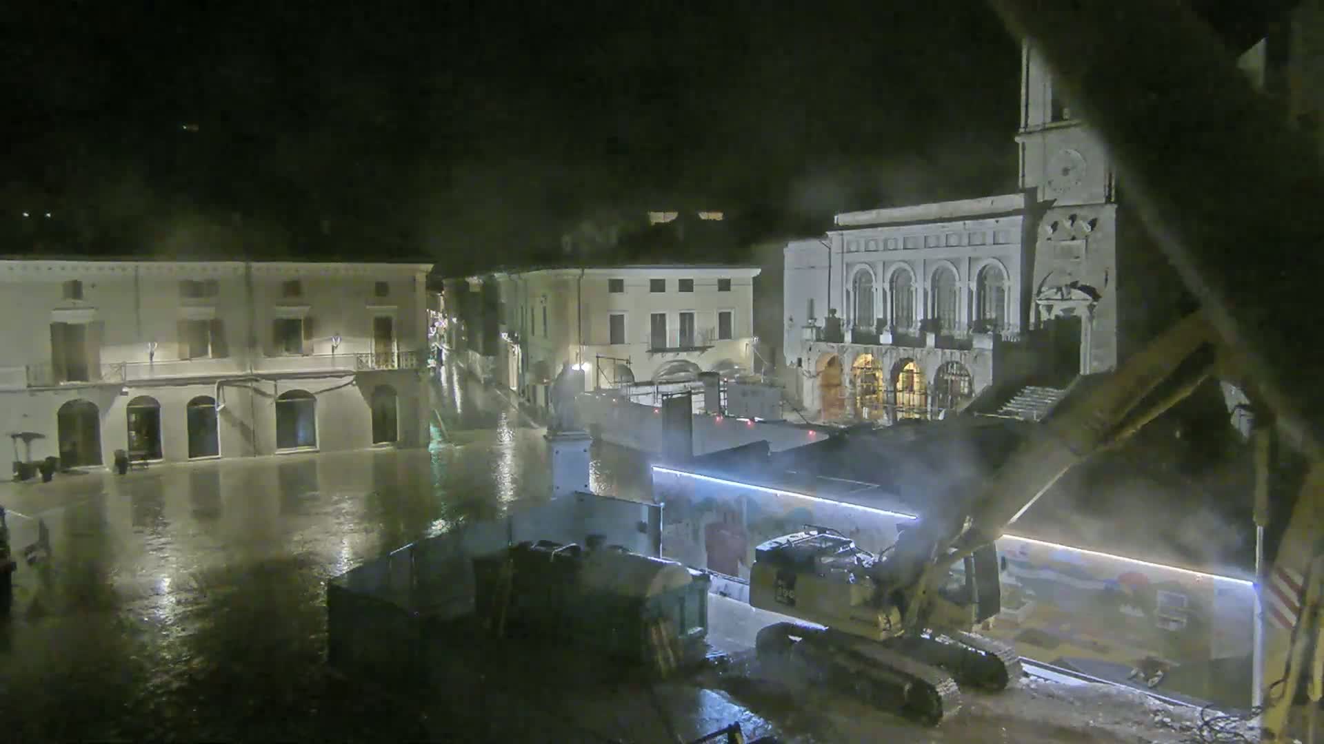 A flooded town square at night is being worked on by construction equipment near several buildings.