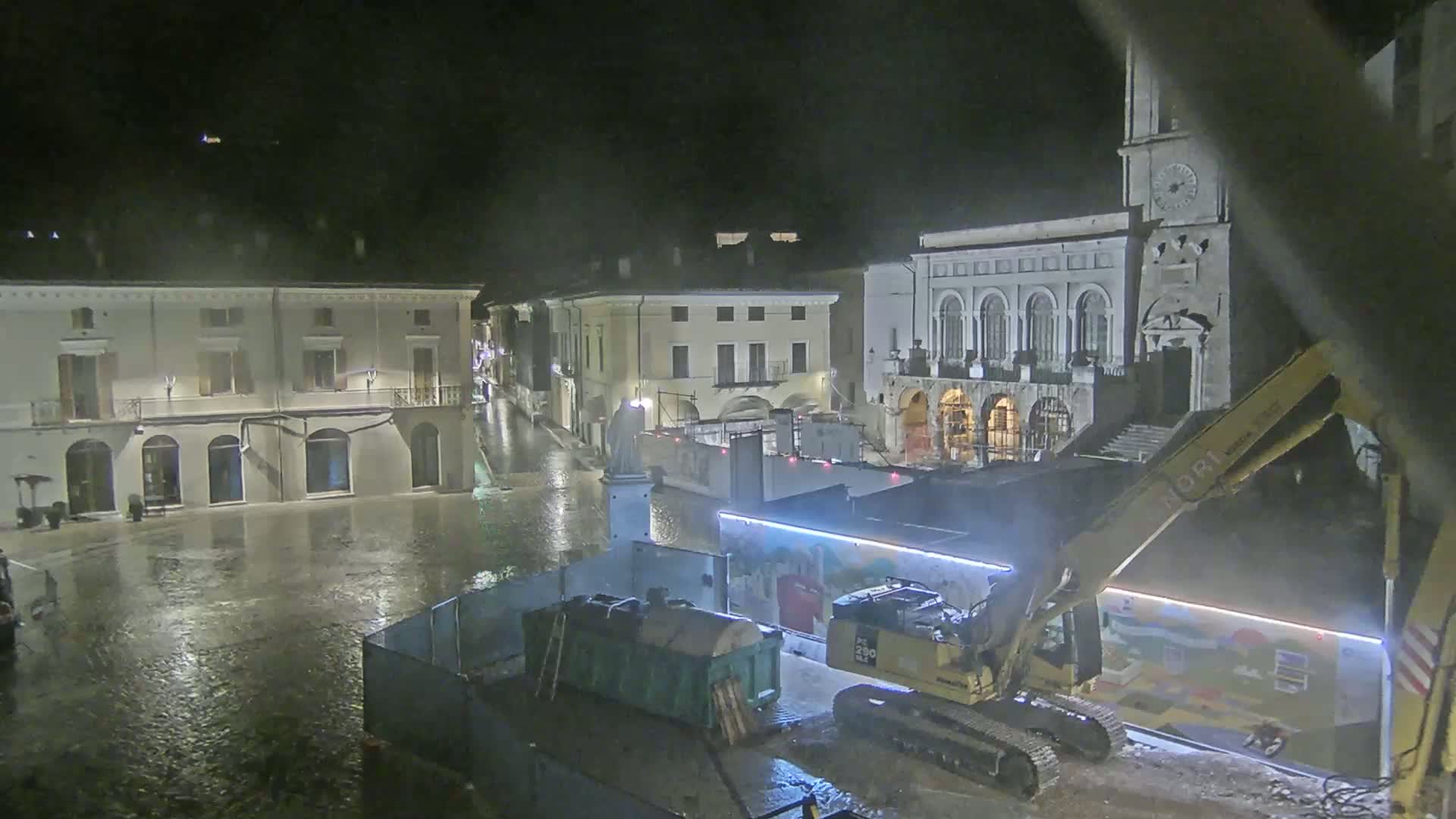 A rain-soaked town square at night is under construction, with an excavator and other equipment present near several buildings.