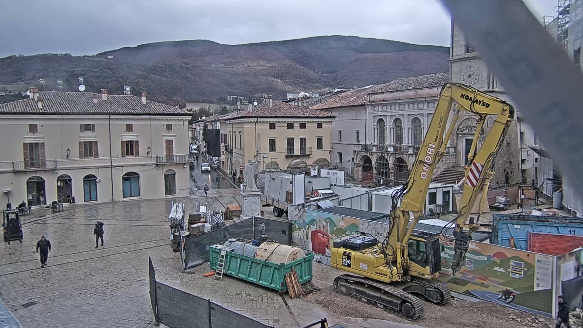 A town square under an overcast sky is undergoing construction, with a large excavator positioned near several shipping containers and other construction equipment.
