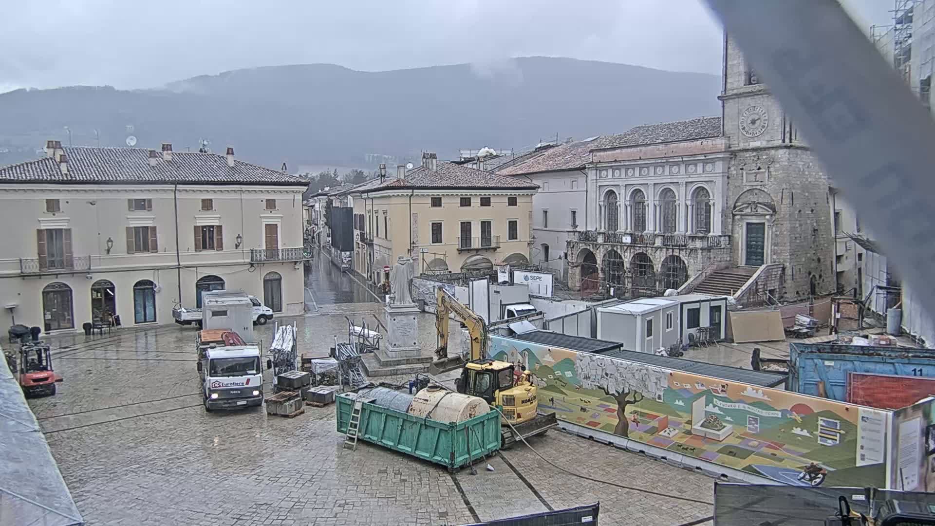 A town square under a gray, overcast sky is undergoing construction, with several pieces of heavy machinery and temporary structures present.