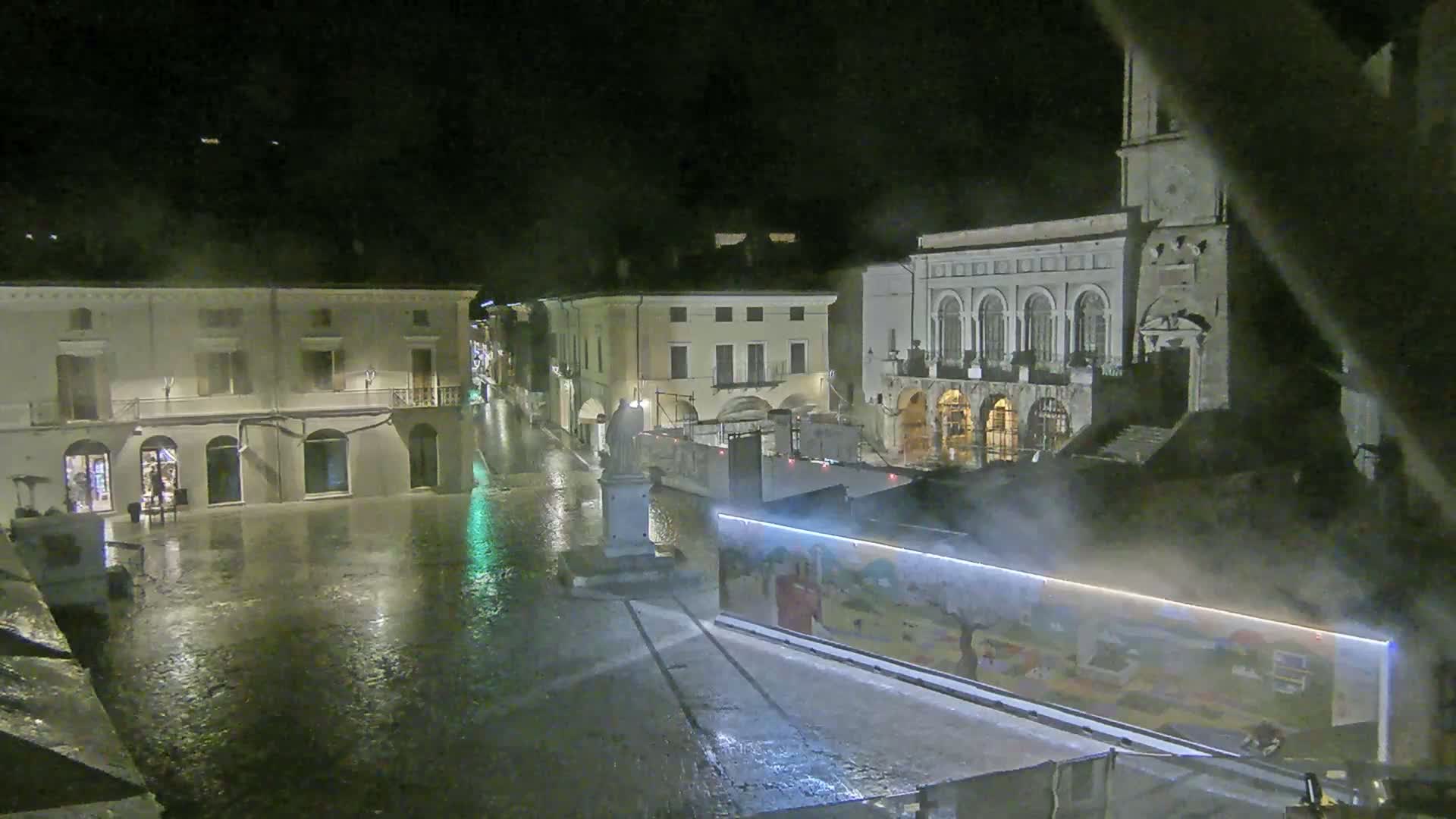 A rain-soaked town square at night is illuminated, showing several buildings, a statue, and a large mural-covered construction barrier.