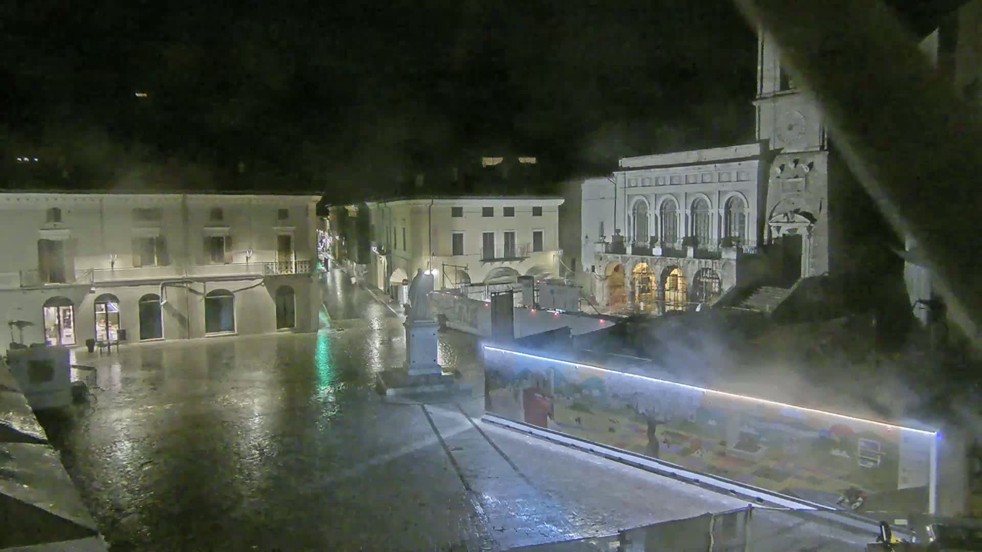 A rain-soaked town square at night, showing several buildings, a statue, and a large construction wall with artwork.