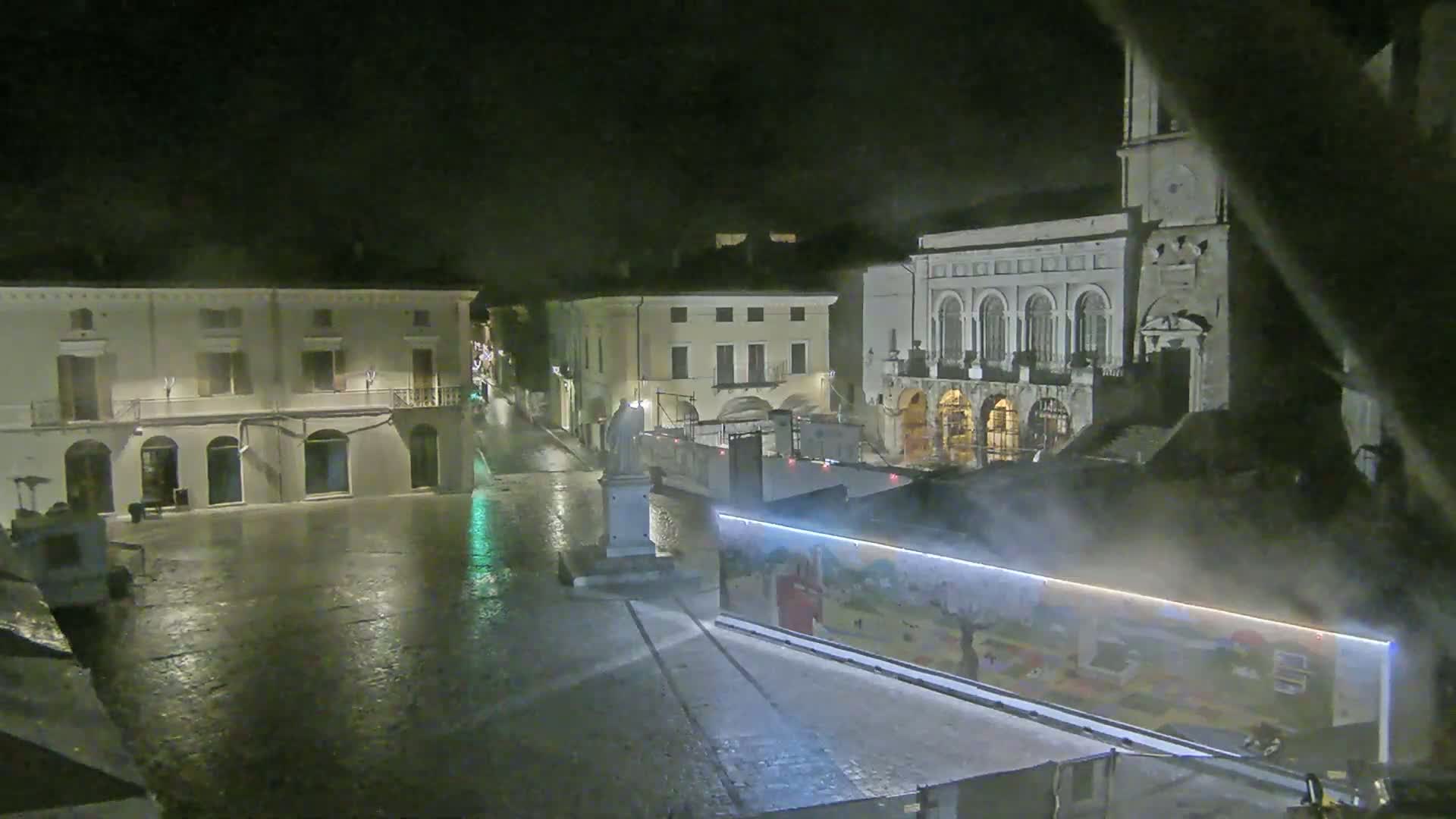 A rain-slicked town square at night, showing several buildings, a statue, and construction barriers with artwork painted on them.