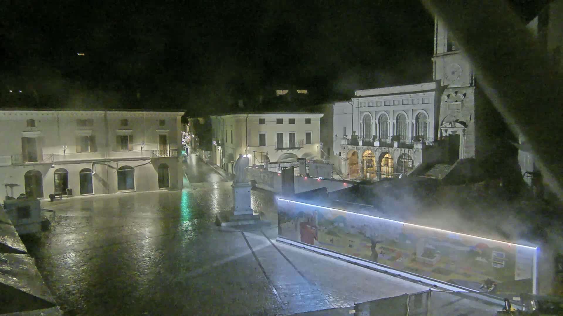 A rain-slicked town square at night, surrounded by buildings, with a large painted wall partially obscuring the view.