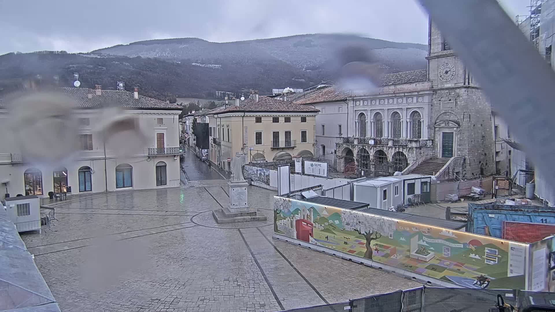 A rain-slicked town square, partially under construction, sits before a backdrop of snow-dusted mountains.