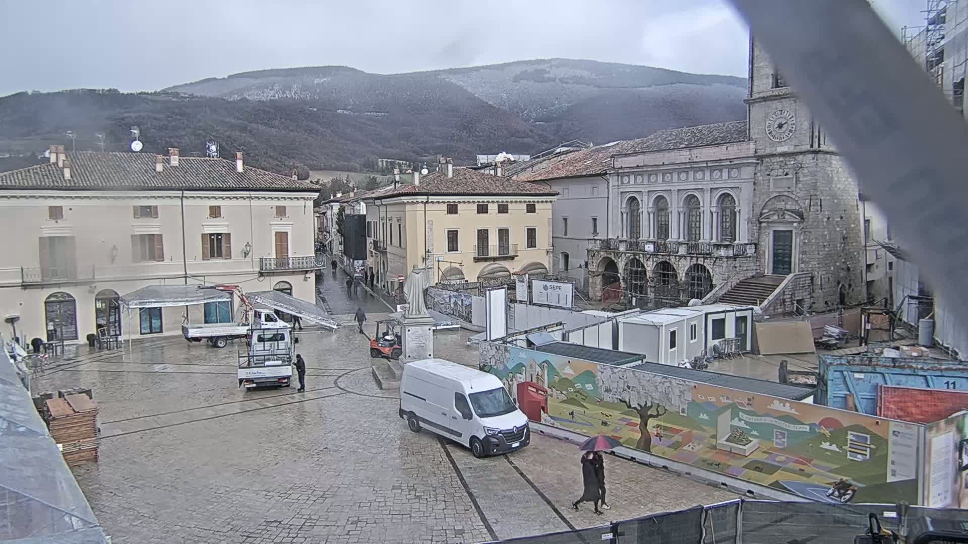 A town square in a mountainous region on a rainy day, with several vehicles, construction materials, and temporary structures present.