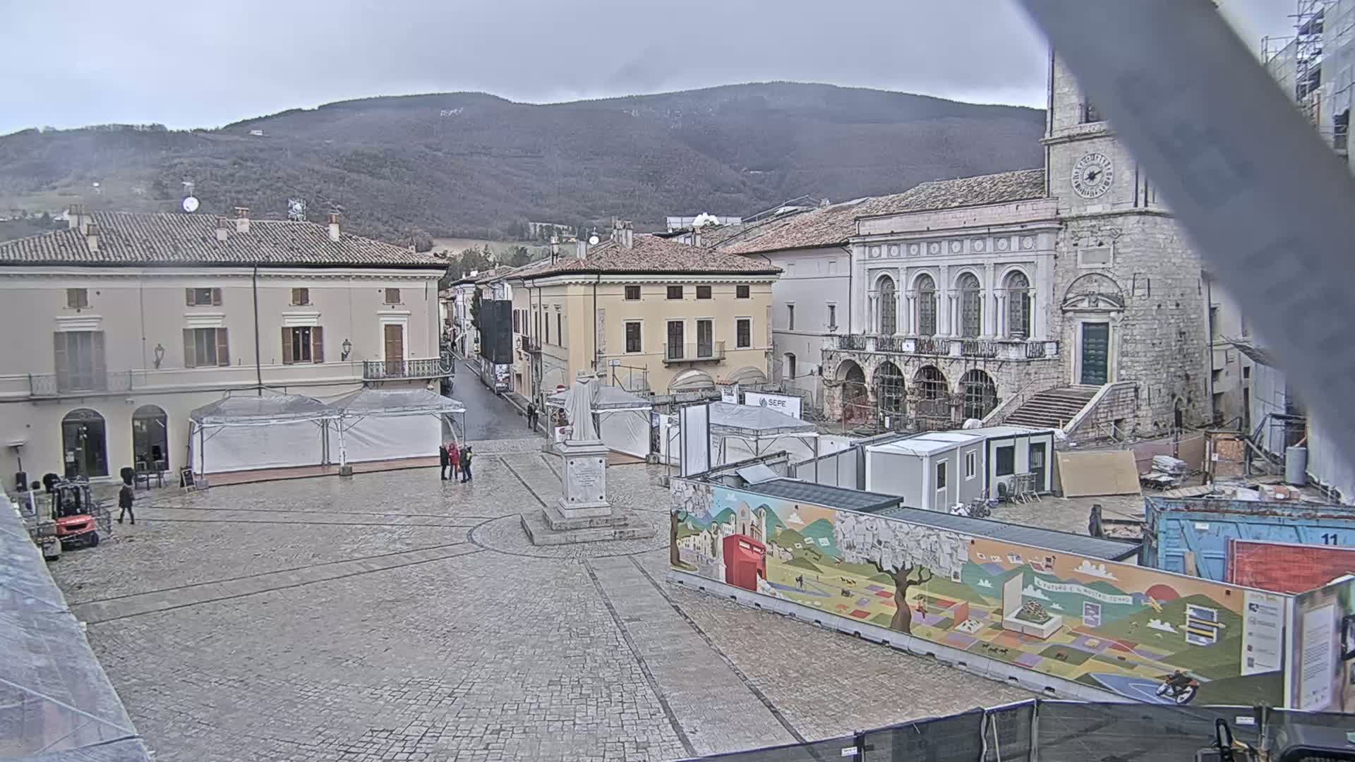A town square under an overcast sky, partially obstructed by construction and temporary structures, is nestled against a backdrop of rolling hills.