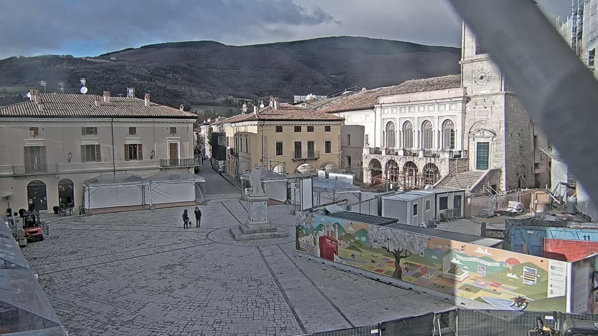 A large town square under an overcast sky is partially obstructed by construction materials and temporary structures, with several buildings surrounding it and mountains visible in the background.