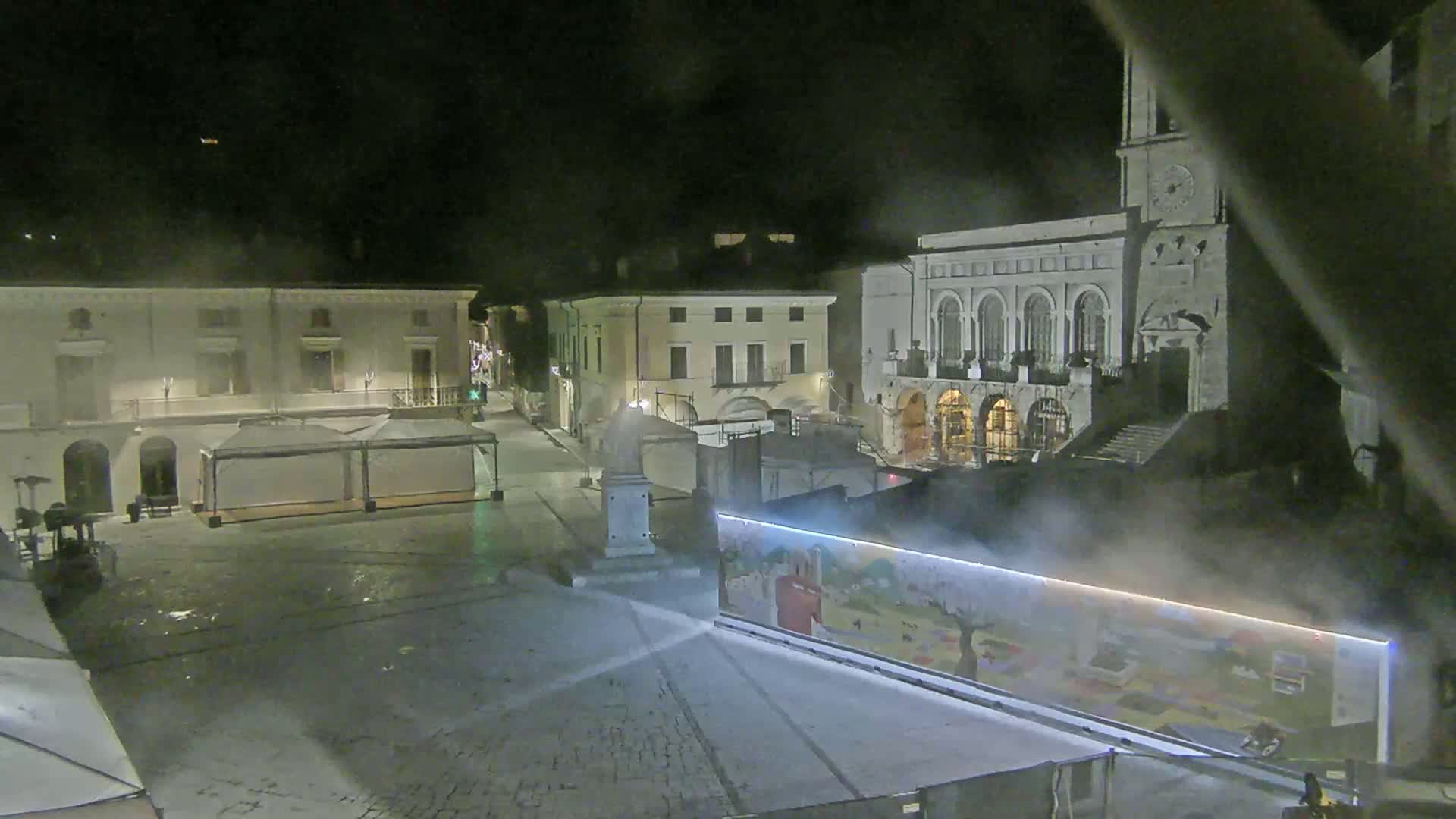 A nighttime view of a town square with several buildings, some tents, and a large mural-covered structure, with a light fog or mist in the air.