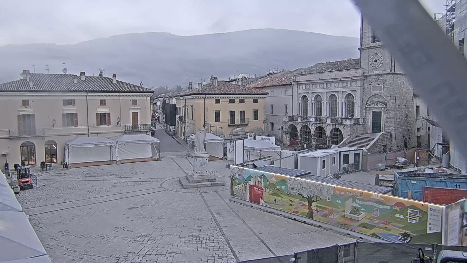 A town square with several buildings, a statue, temporary structures, and construction materials under an overcast sky.