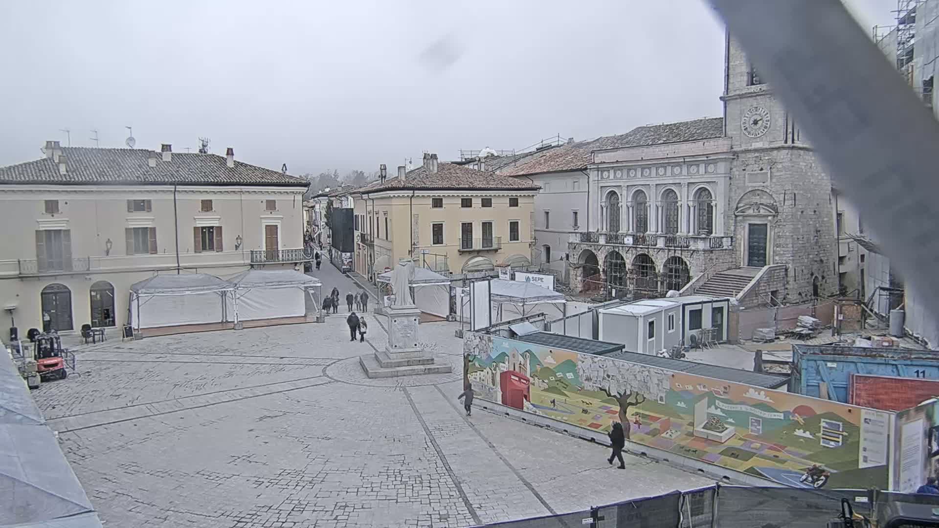 A mostly empty town square, under an overcast sky, is partially surrounded by historic buildings and temporary structures, including tents, construction barriers, and shipping containers decorated with murals.