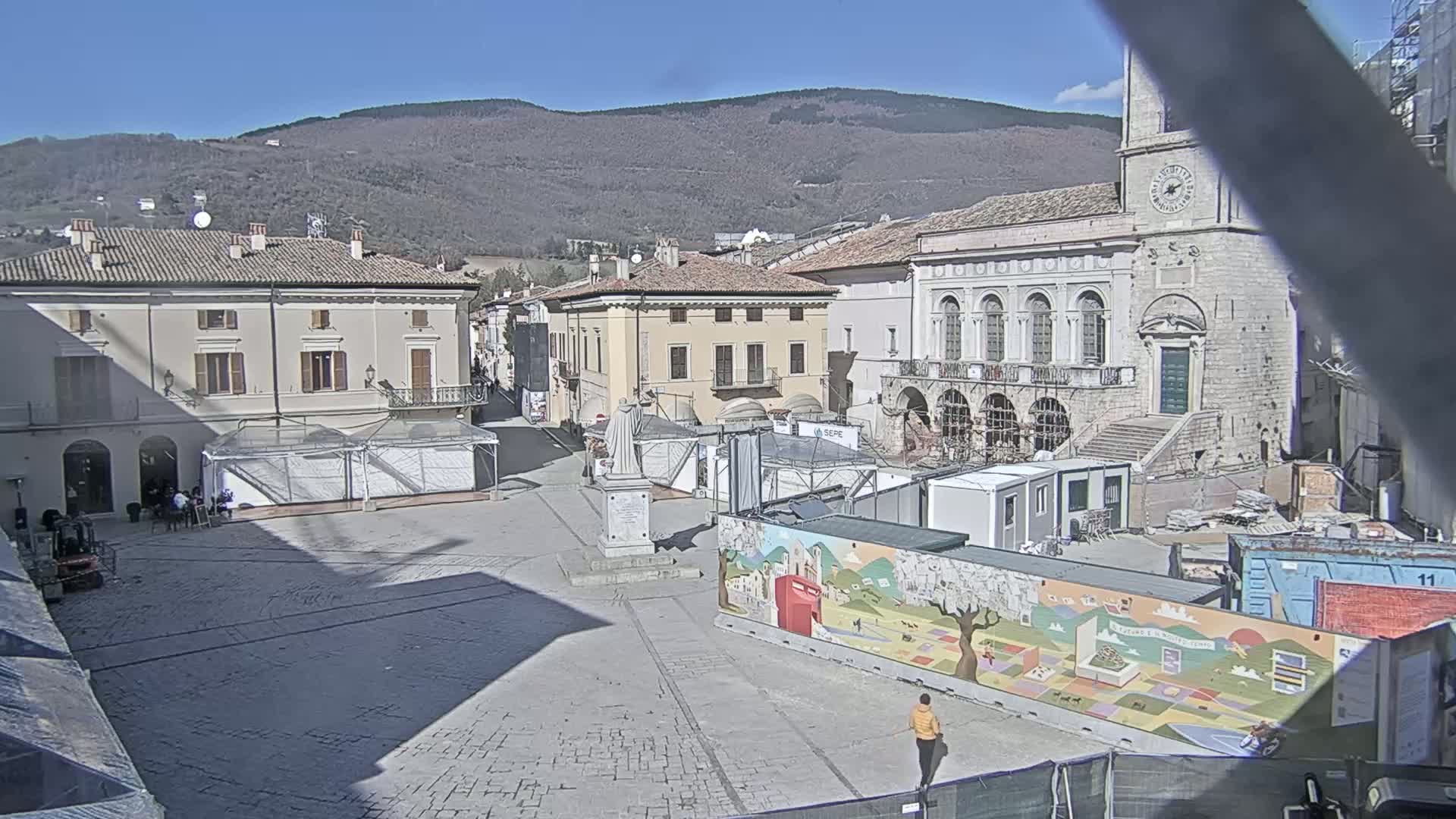 A mostly empty town square, under a clear sunny sky, is surrounded by buildings and currently undergoing construction or renovation.