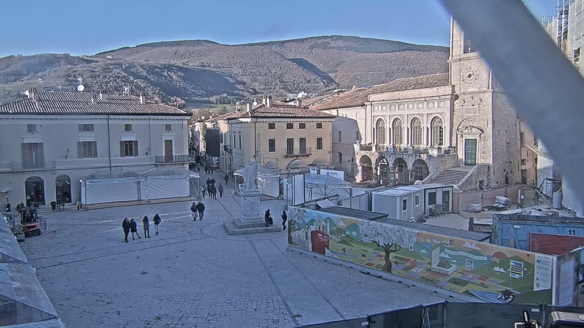 A town square with several buildings, some under construction, is seen on a sunny day with mountains in the background.