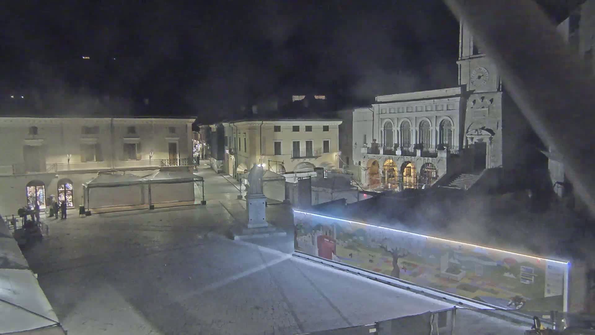 A nighttime view of a town square with several buildings, including a large one with arched windows, some temporary market stalls, and a statue, all under a hazy or foggy atmosphere.