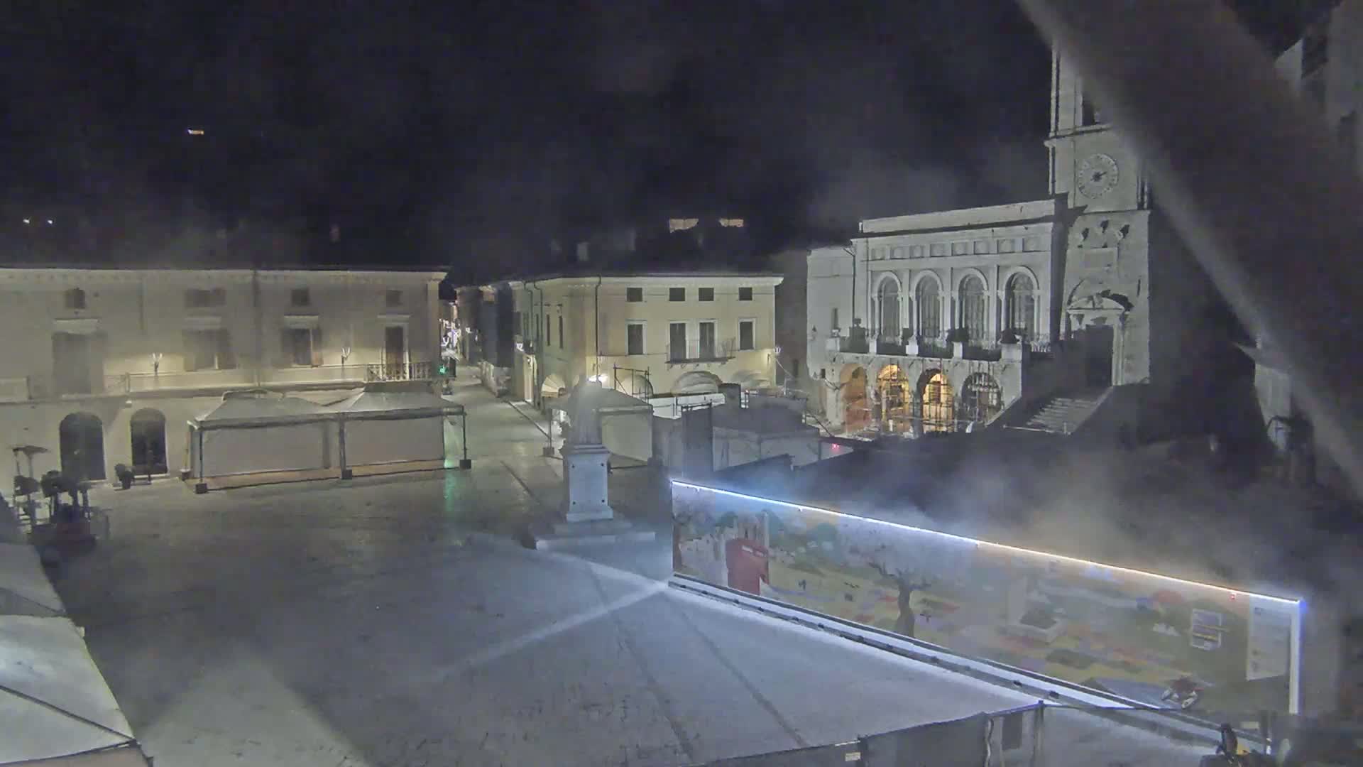 A nighttime view of a town square with several buildings, temporary structures, and a large painted mural, under hazy conditions.