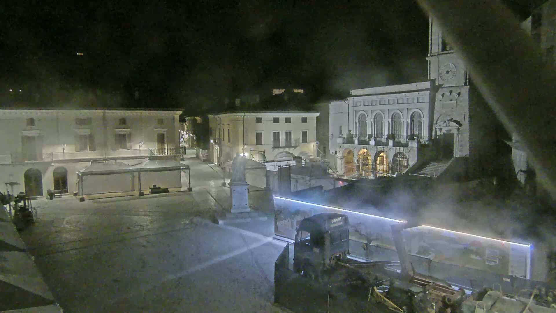 A nighttime view of a town square with several buildings, including a large structure with arched windows and a clock tower, partially obscured by fog or mist.