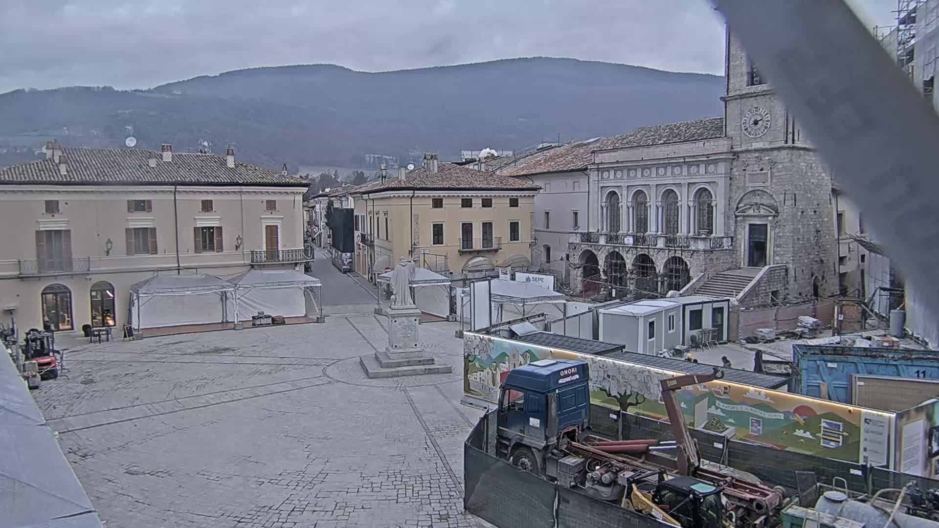 A town square under an overcast sky is partially obstructed by construction materials and equipment, with several buildings and a statue visible in the background.